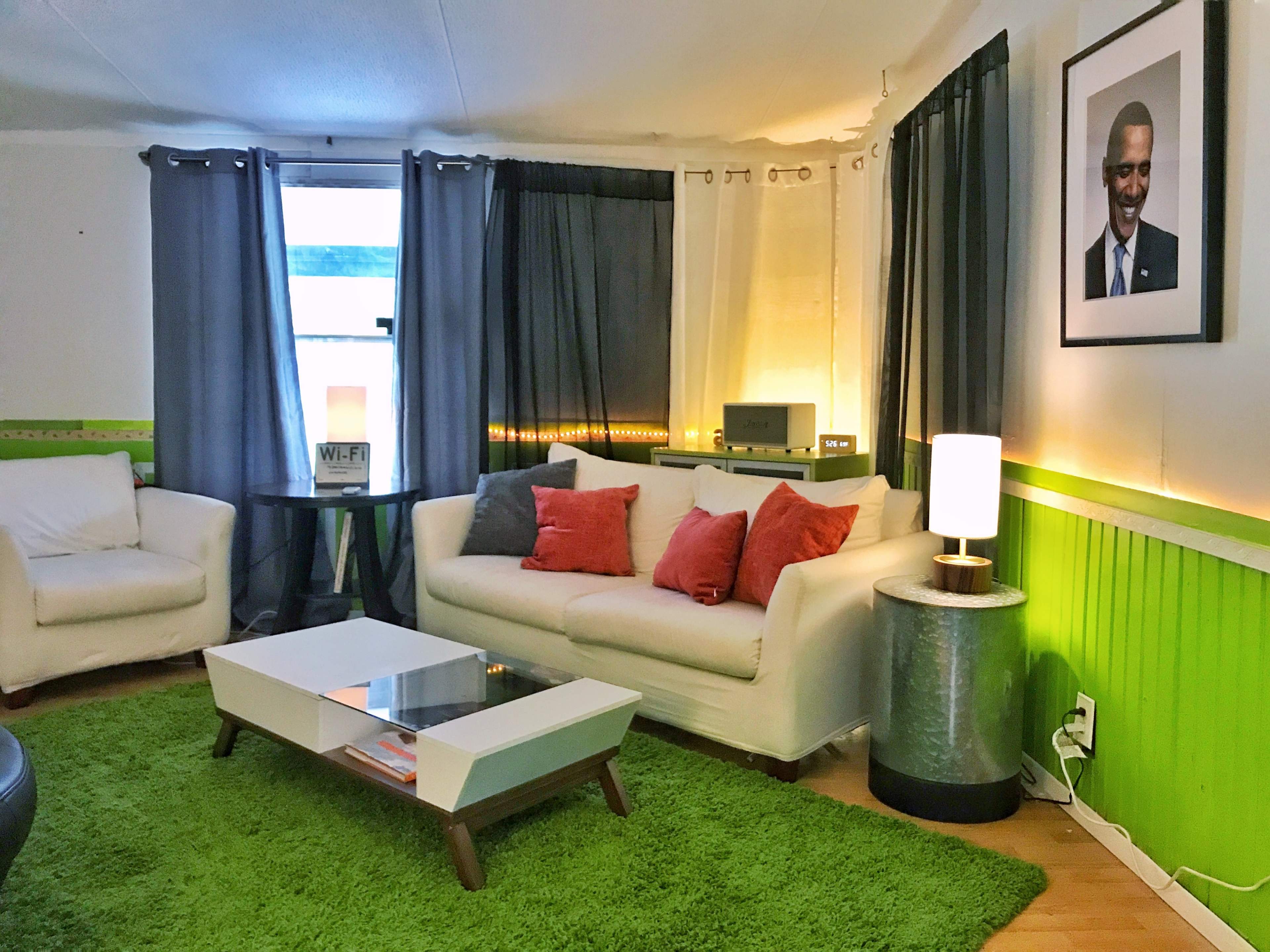 A modern living room features a white sofa with colorful pillows, a green area rug, a coffee table, and a portrait on the wall alongside curtains and lighting.