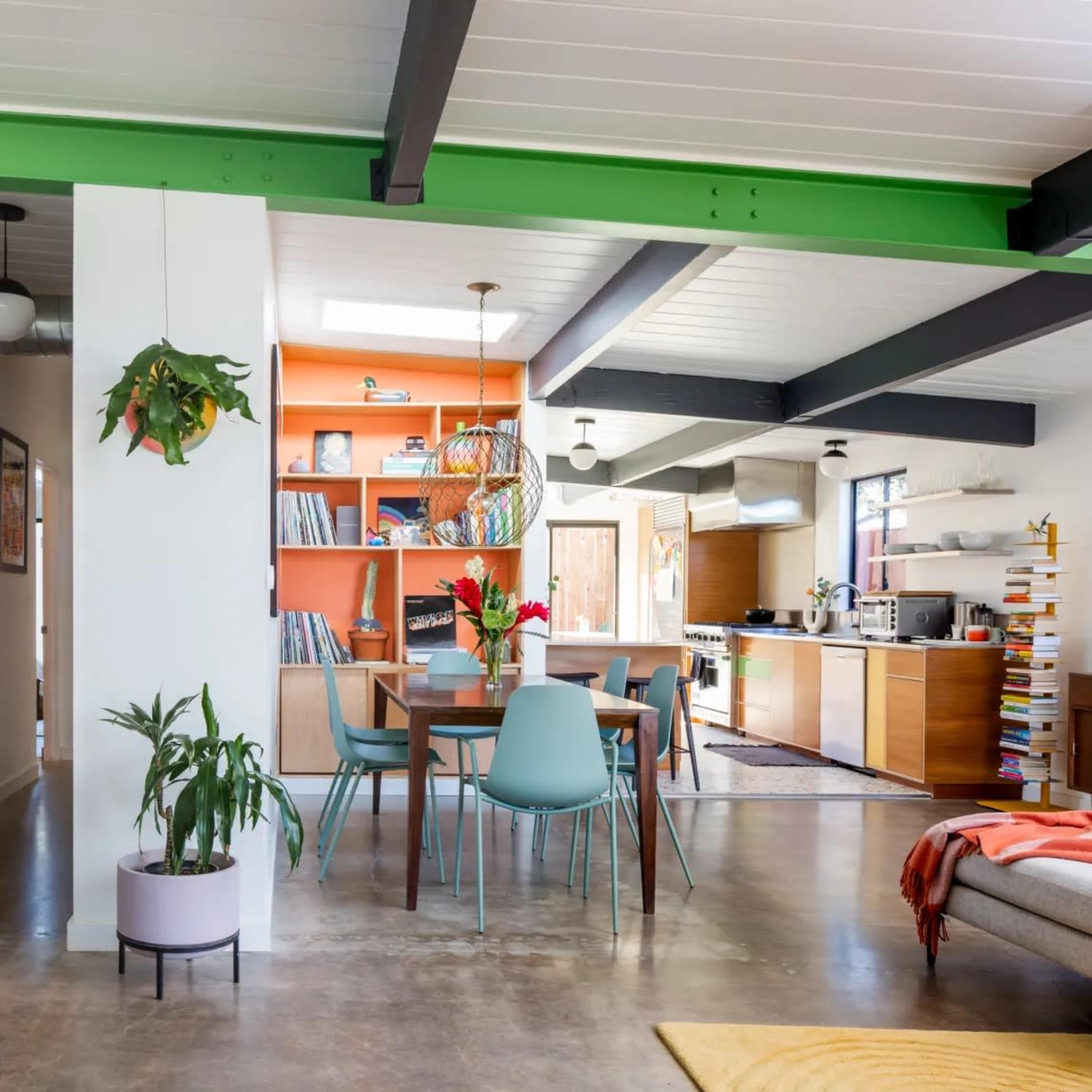 A modern, open-concept living space featuring a dining area with a table and chairs, a bookshelf, and a kitchen in the background.