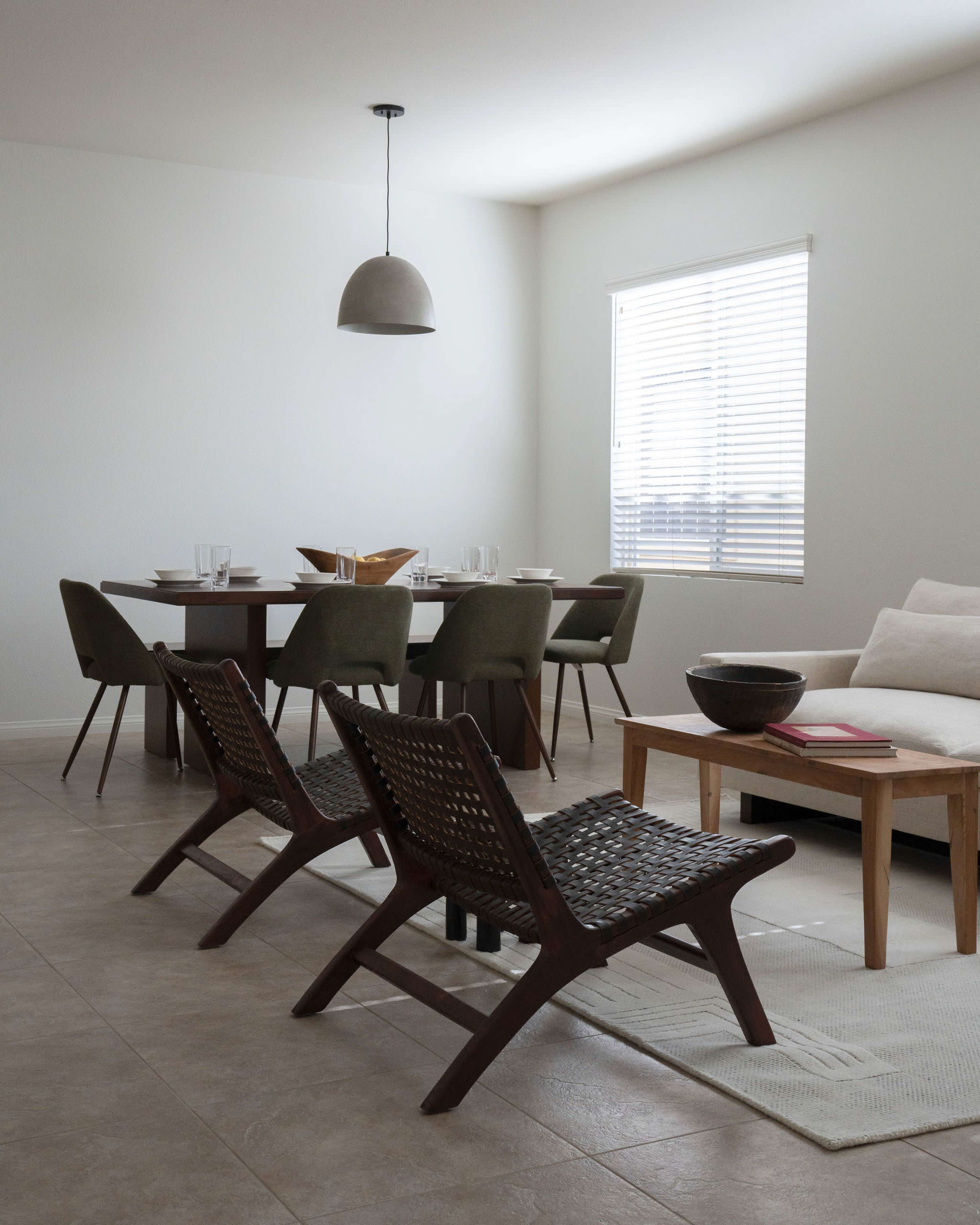 A modern, minimalistic living space features a dining table with chairs, two woven lounge chairs, and a sofa, all arranged on tiled flooring.