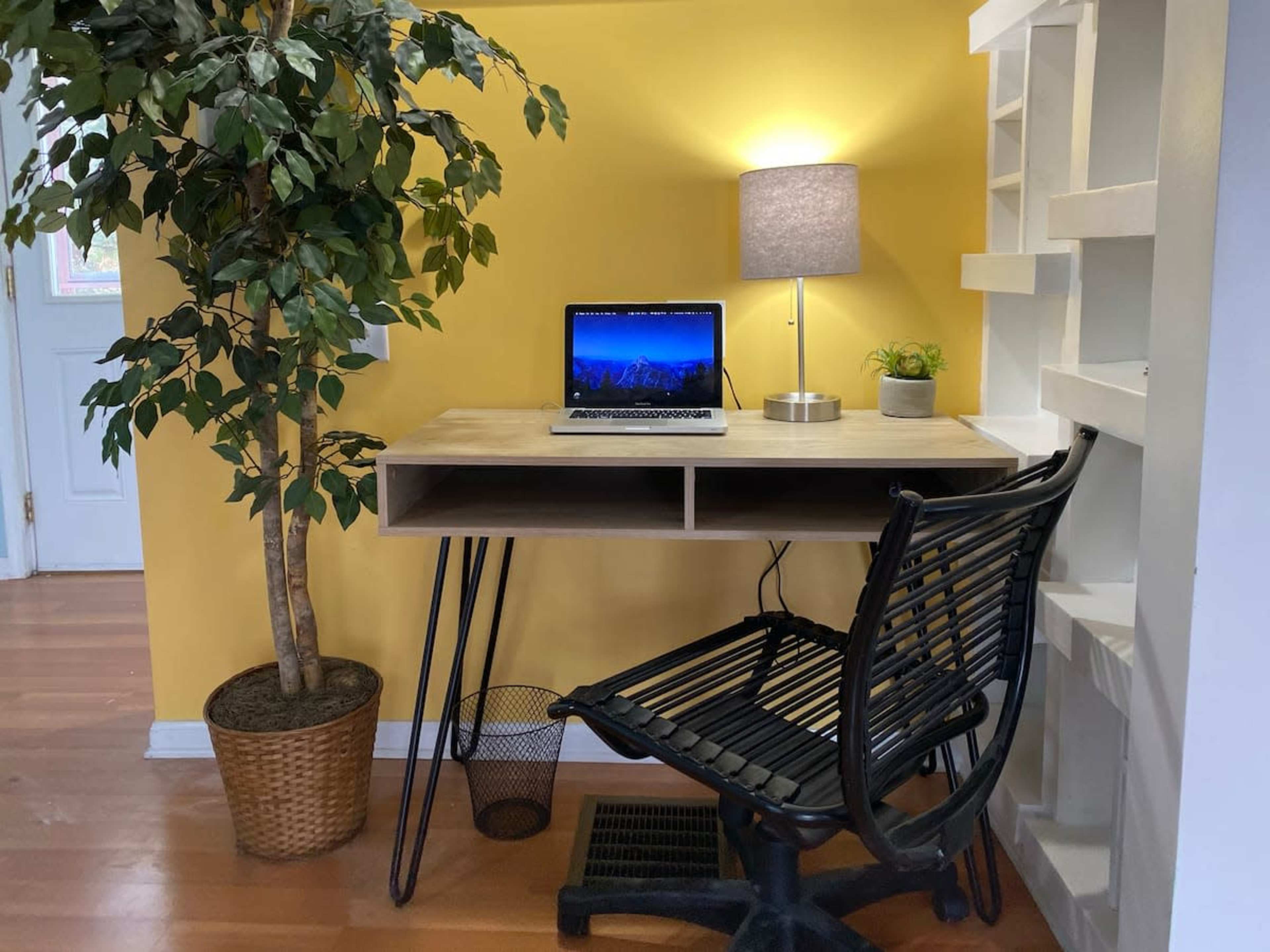 A small workspace features a wooden desk with a laptop, a lamp, a potted plant, and a black chair, set against a yellow wall next to a decorative tree.