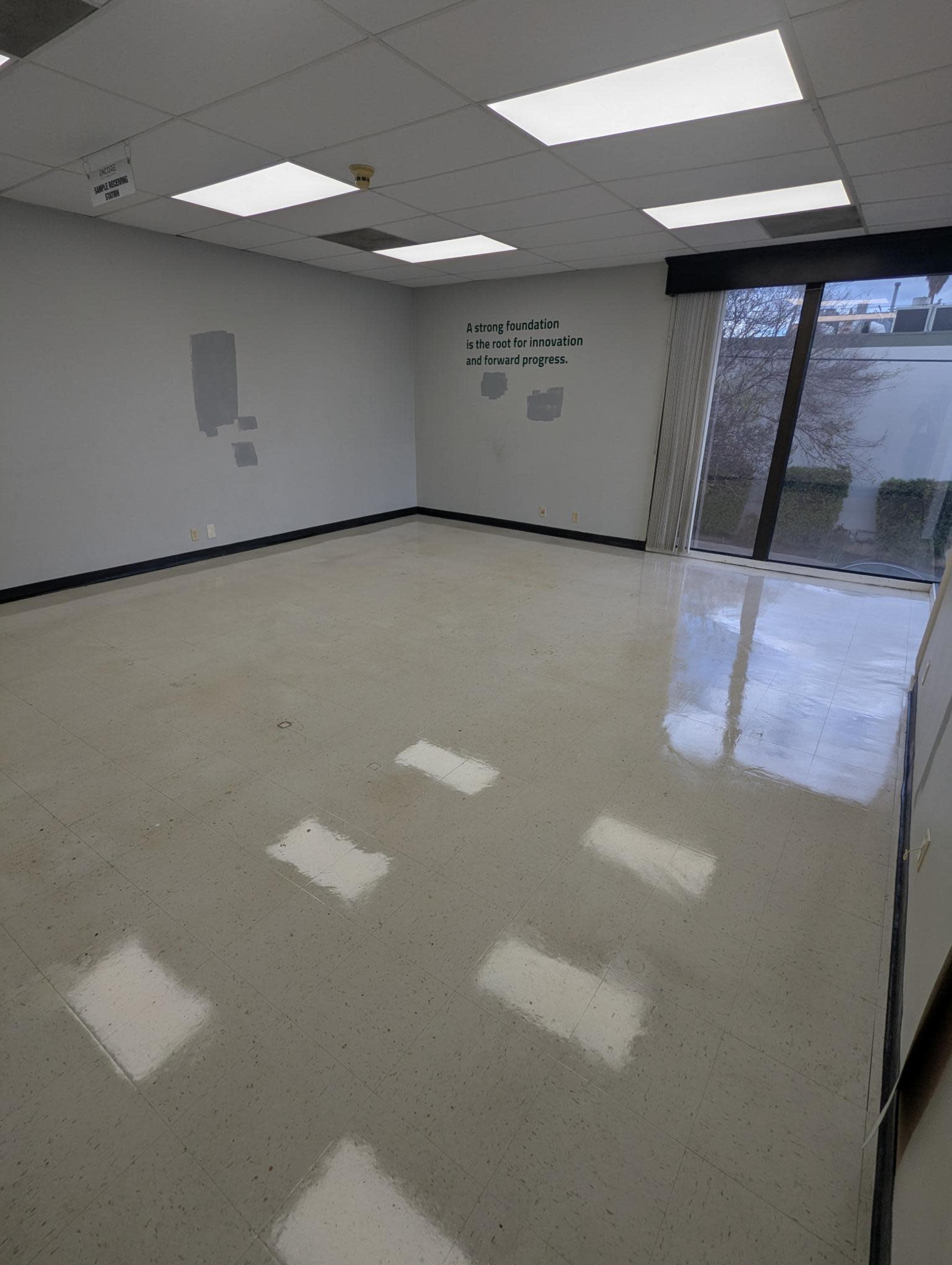 The image shows a vacant, polished room with tiled floors and gray walls, featuring a large window and wall text about innovation and progress.