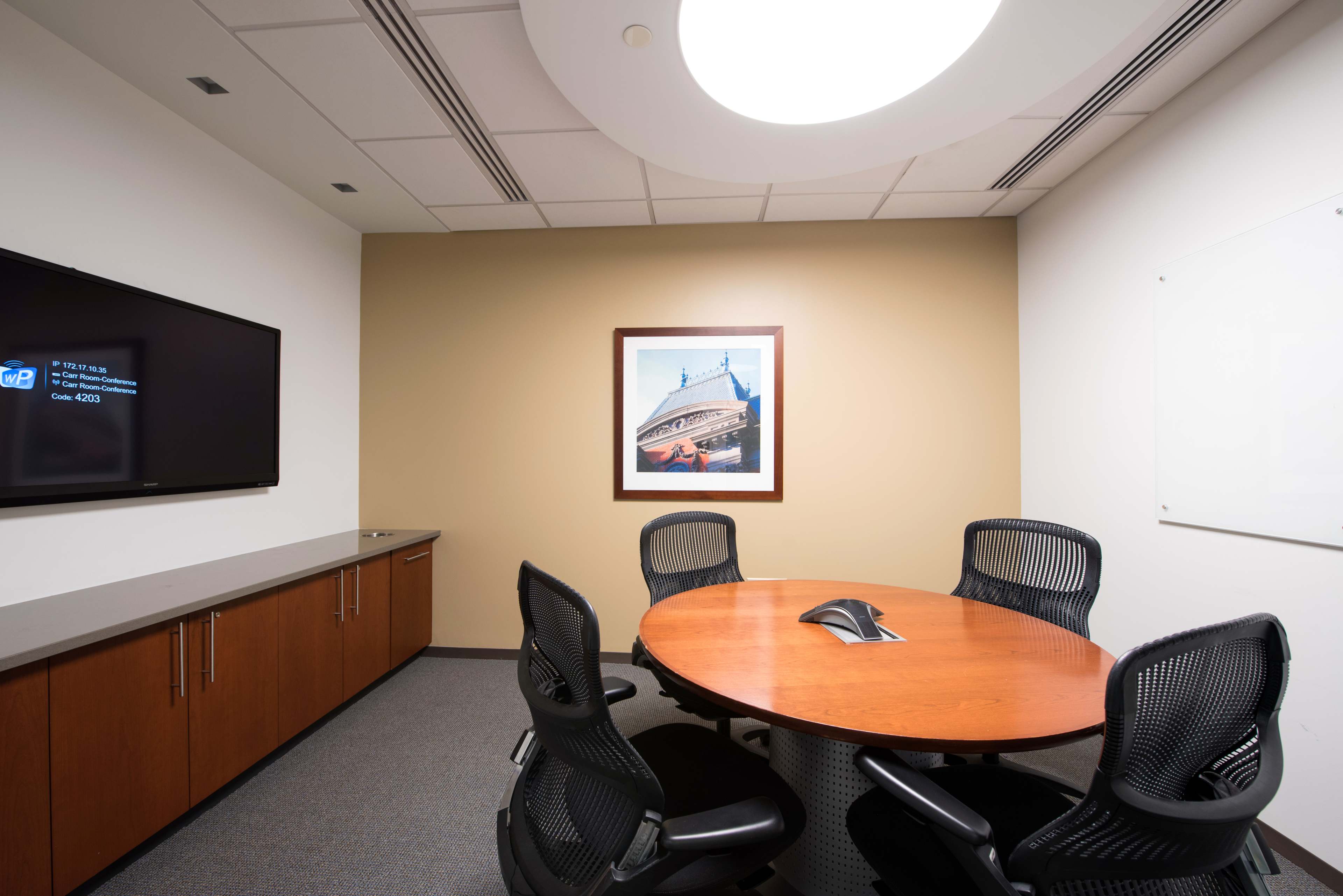 A small conference room with a round wooden table, four black chairs, a wall-mounted television, and a framed picture on the wall.