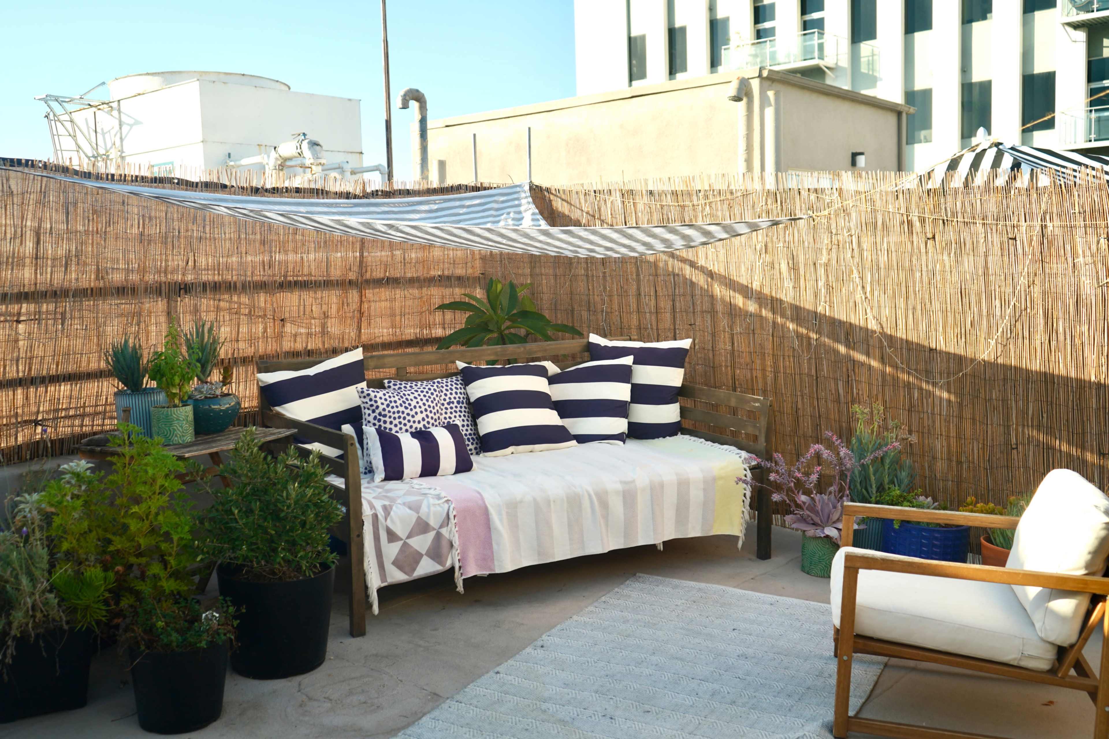 A small outdoor seating area features a wooden sofa adorned with striped cushions, surrounded by potted plants and set against a bamboo privacy screen.