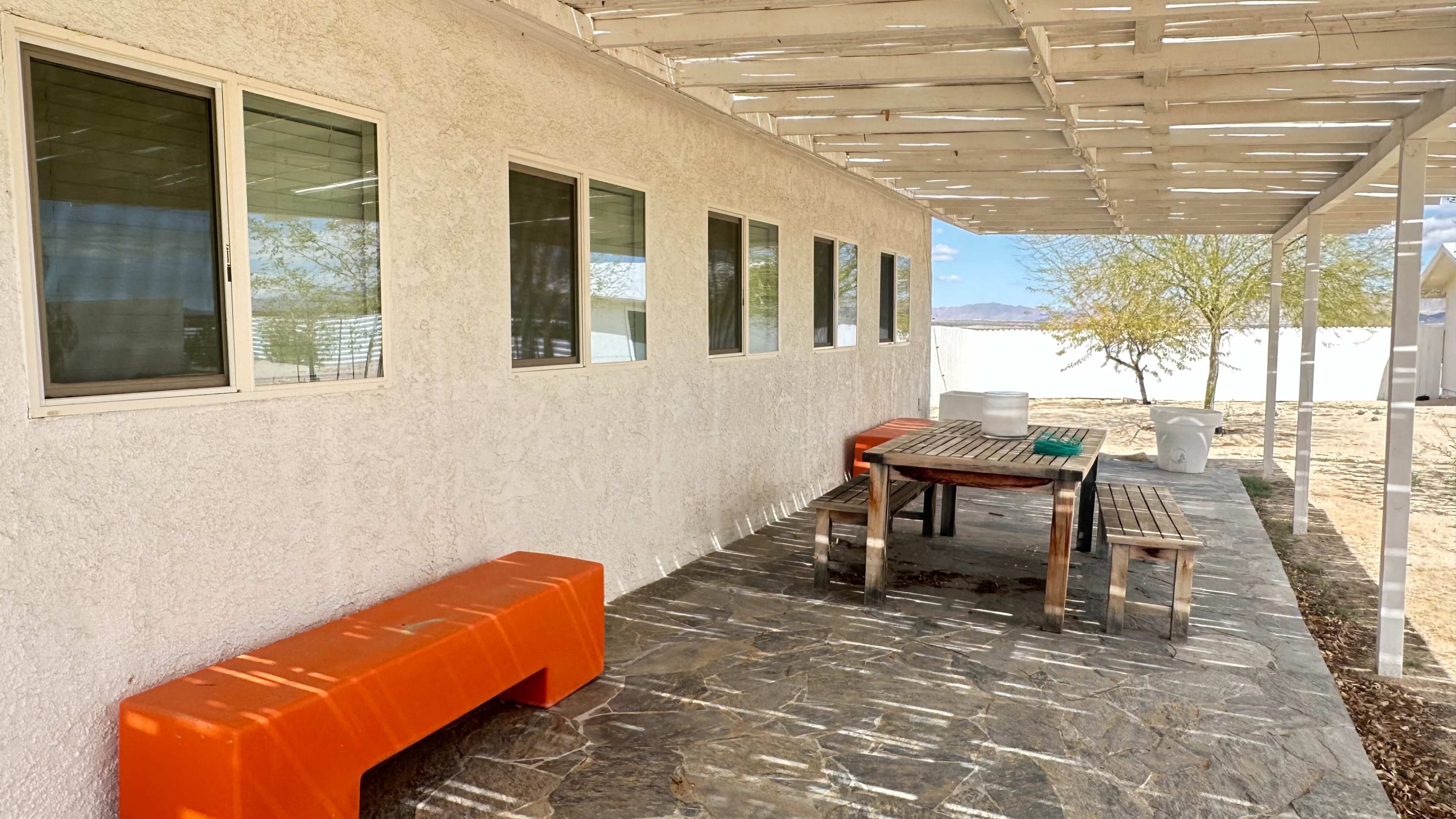 A covered patio features a wooden table and benches on a stone floor, with orange seating and multiple windows along the wall.