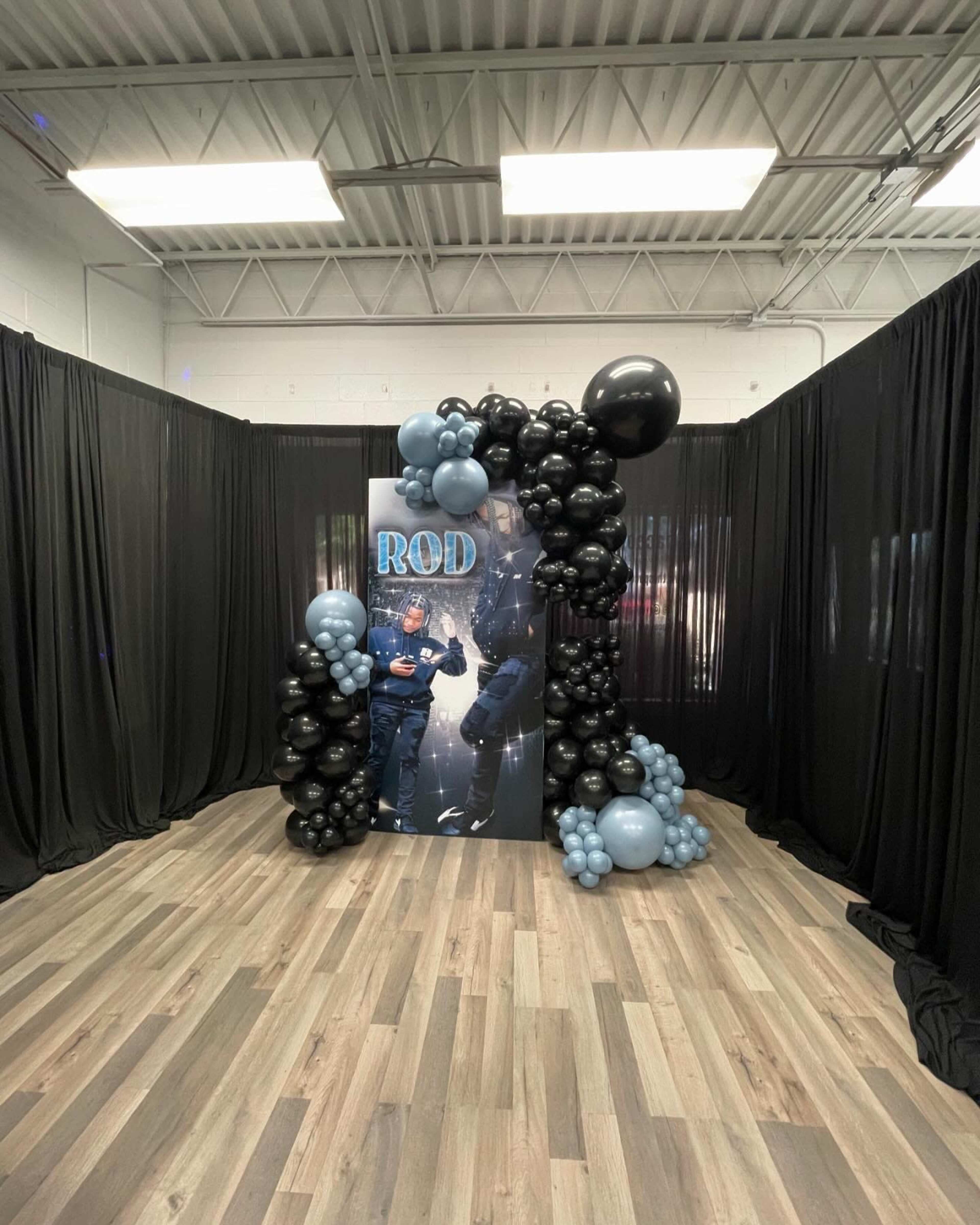 A balloon arch made of black, gray, and blue balloons frames a backdrop featuring a person in a police-like uniform, set against black drapes in a spacious event area.