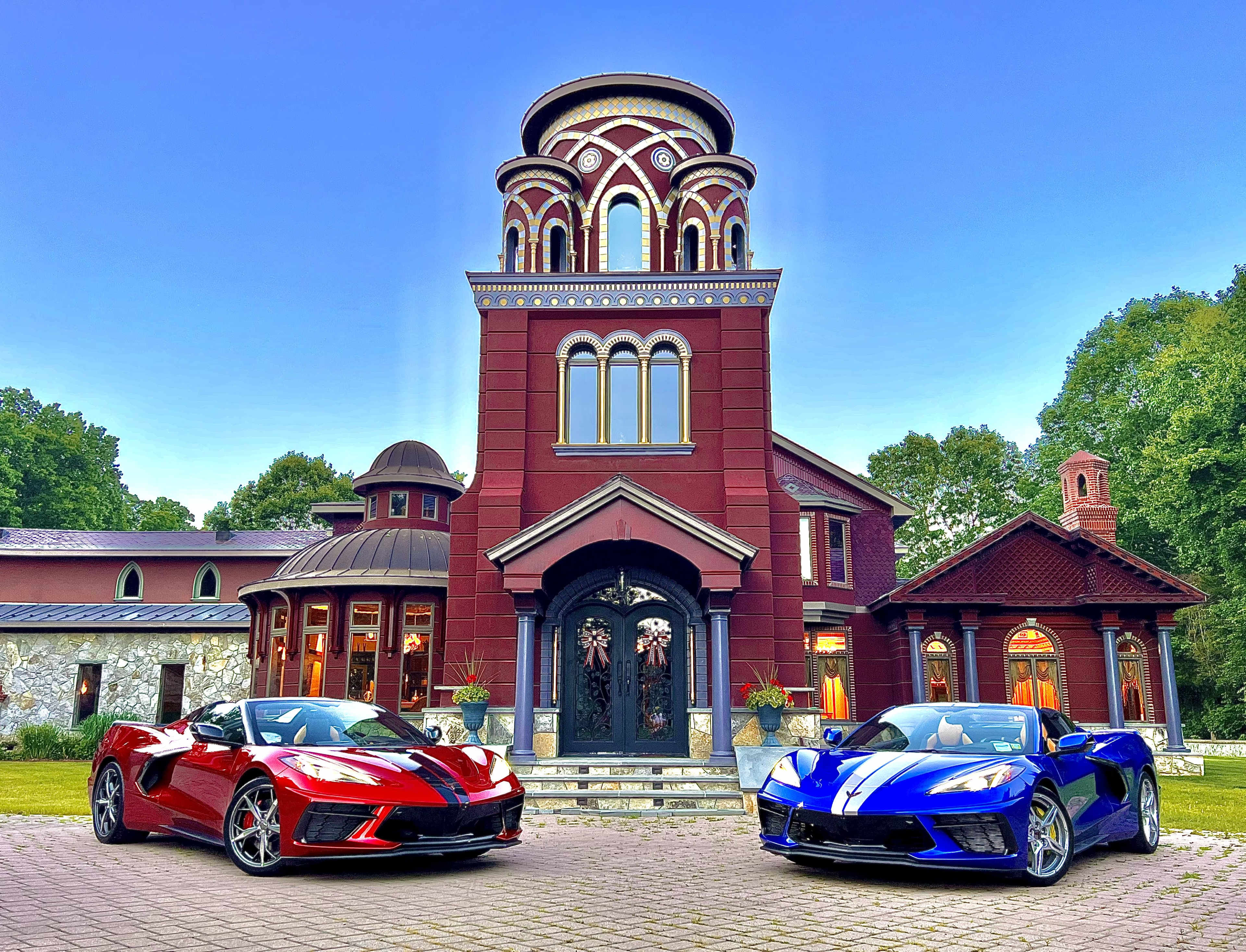 Two Chevrolet Corvette convertibles, one red and one blue, are parked in front of a large red and brick building with unique architectural features and a blue sky in the background.