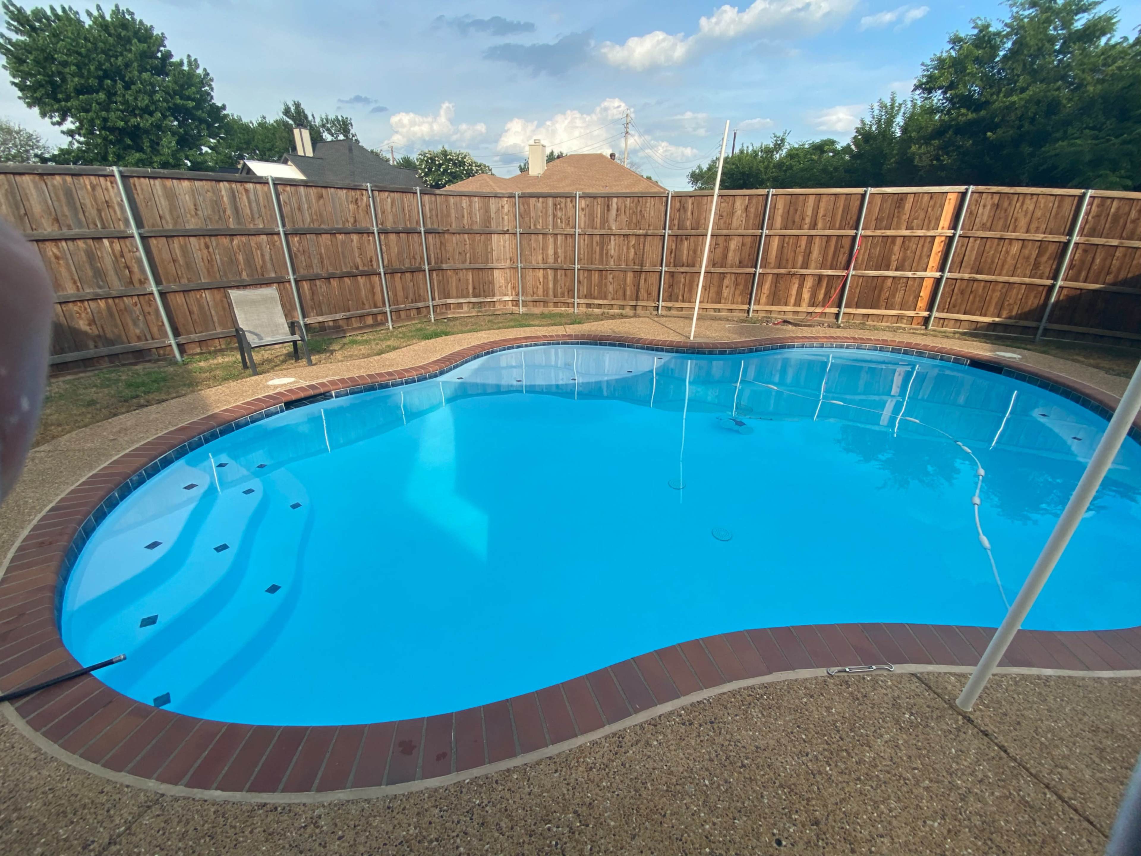 The image shows a residential swimming pool with a curved design, surrounded by a wooden fence and a flat grassy area.