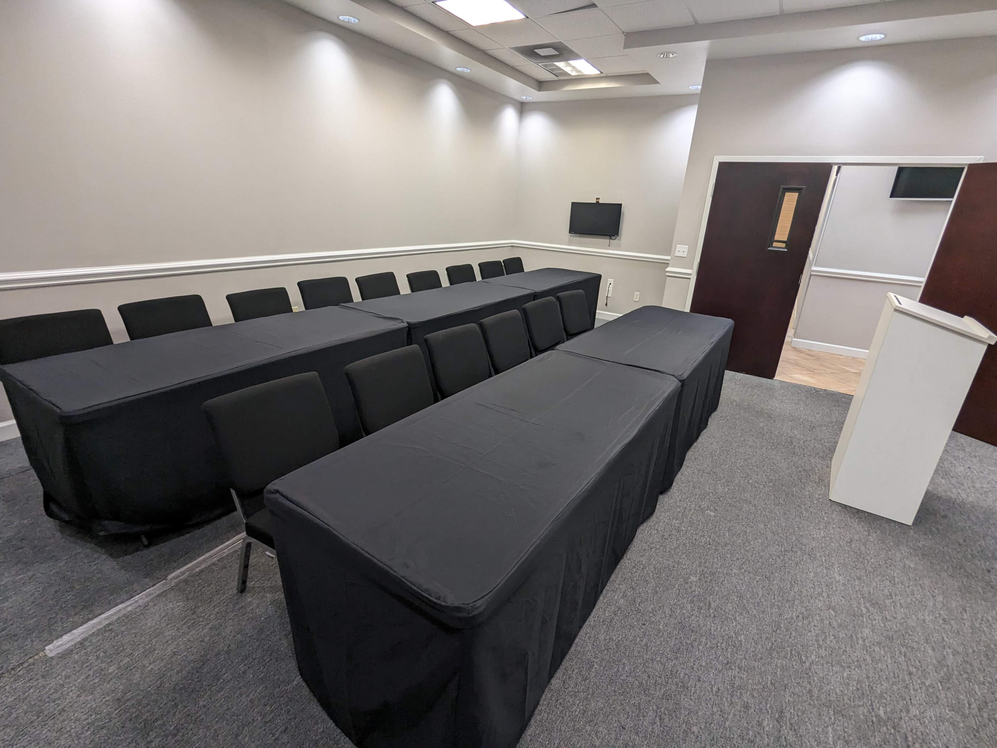 A conference room features multiple rows of black-covered tables and chairs, with a podium at the front and a wall-mounted screen.