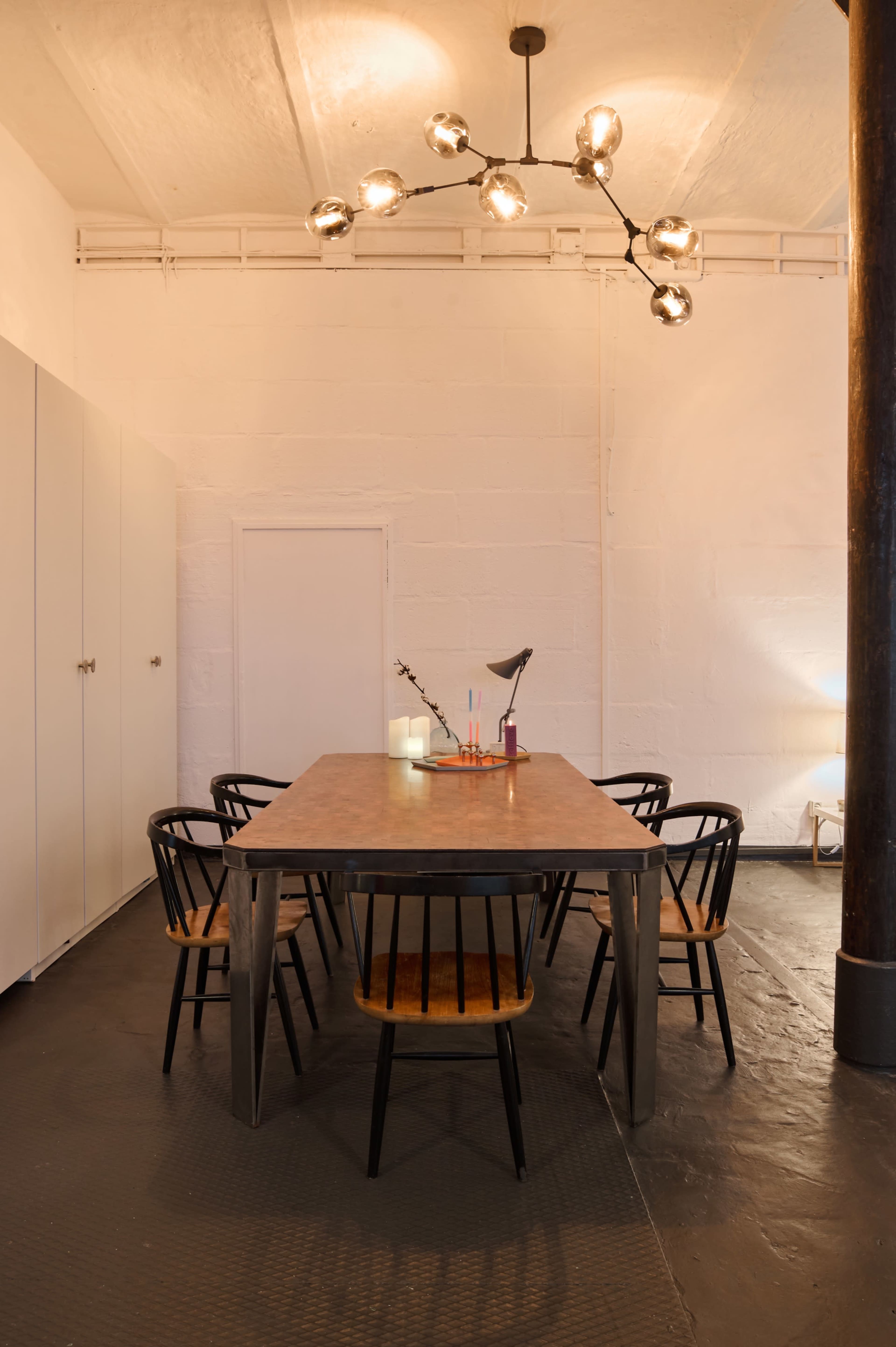 The image shows a minimalist meeting room with a large metal dining table surrounded by black chairs, a modern light fixture, and white walls.