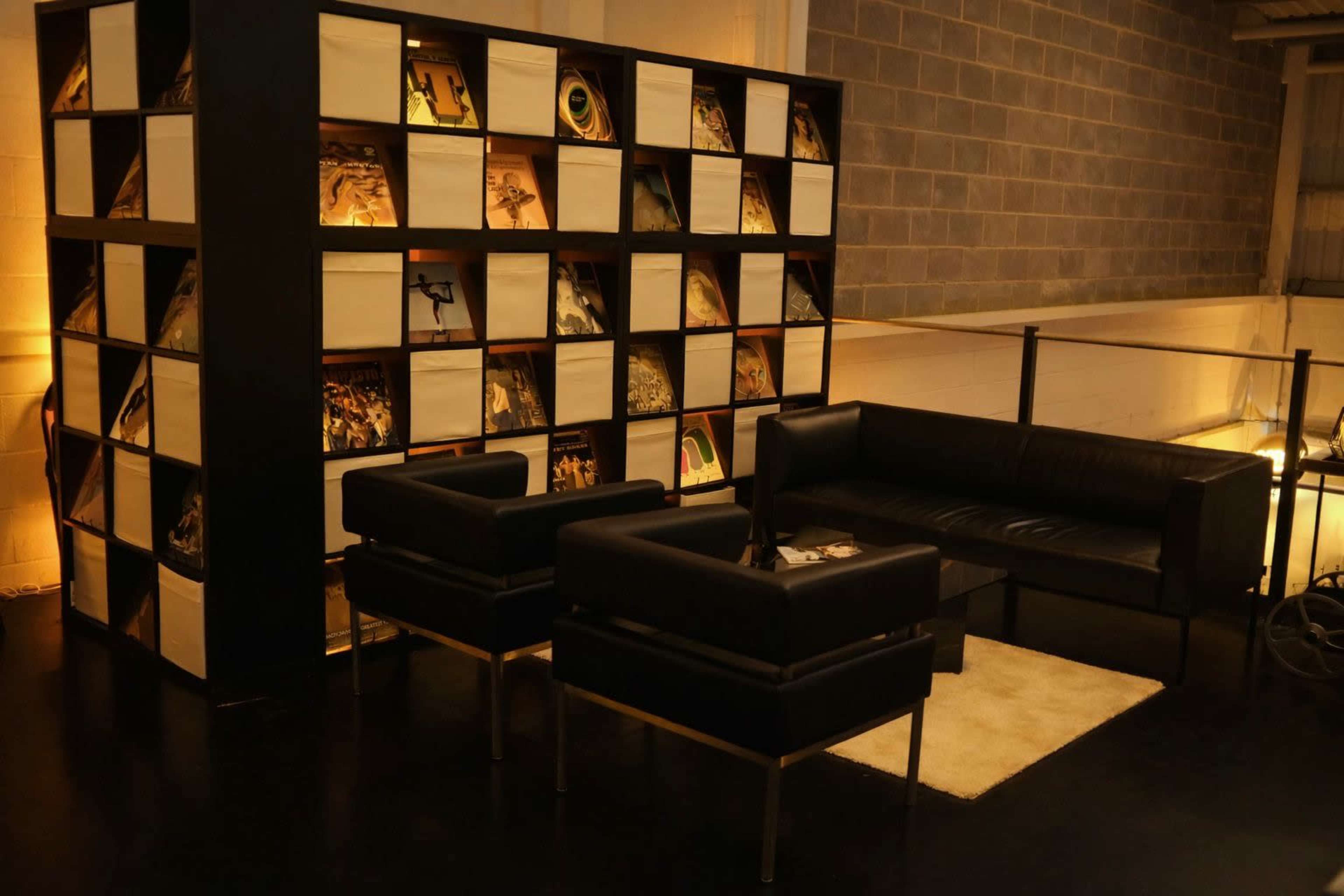 The image shows a modern seating area featuring black chairs and a sofa, with a bookshelf filled with records in the background.