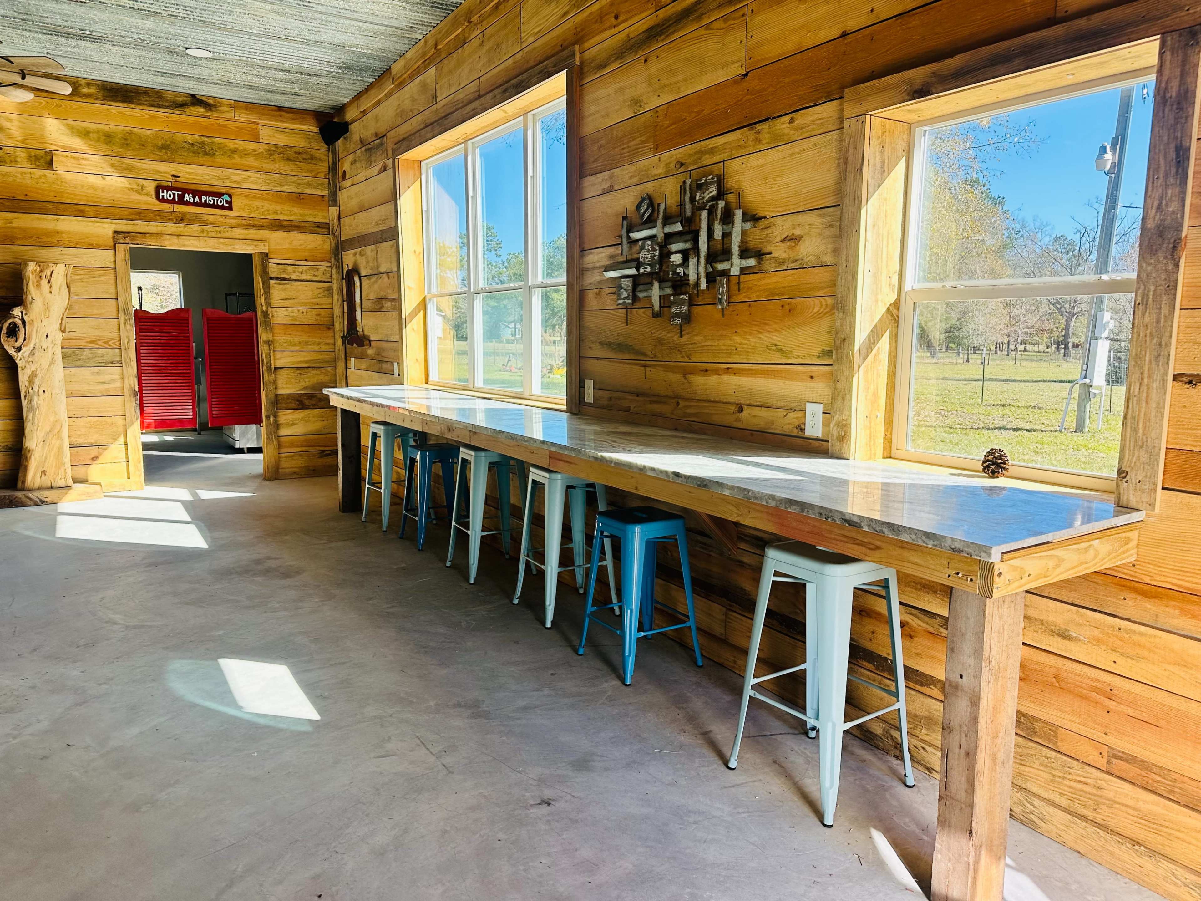 A rustic interior with wooden walls, a long white countertop with blue stools, and large windows allowing natural light to enter.
