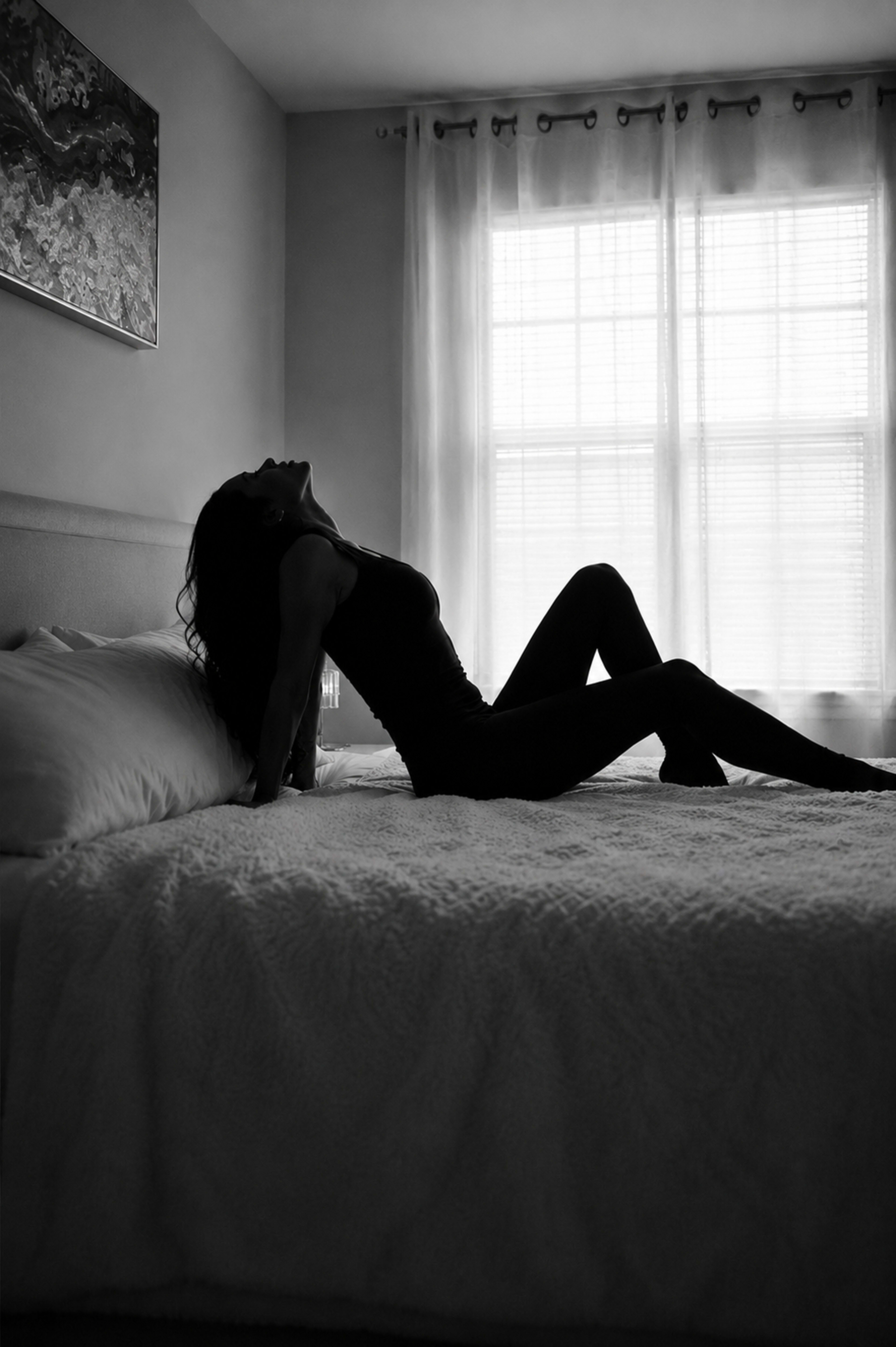 A person is sitting on a bed with their back against the headboard, silhouetted against a window with light streaming through.