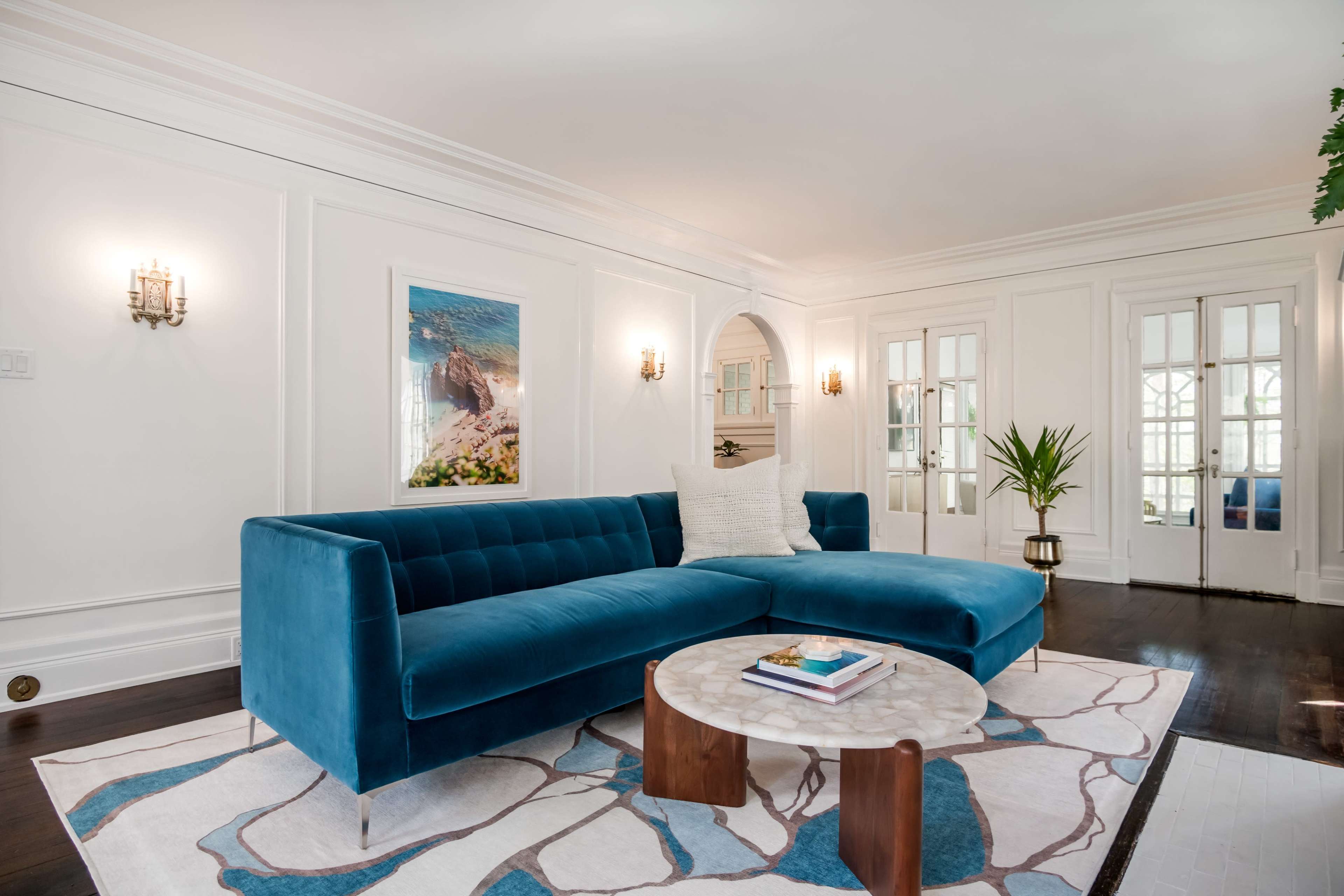 A spacious living room features a blue sectional sofa, a marble coffee table, and large windows allowing natural light to enter.
