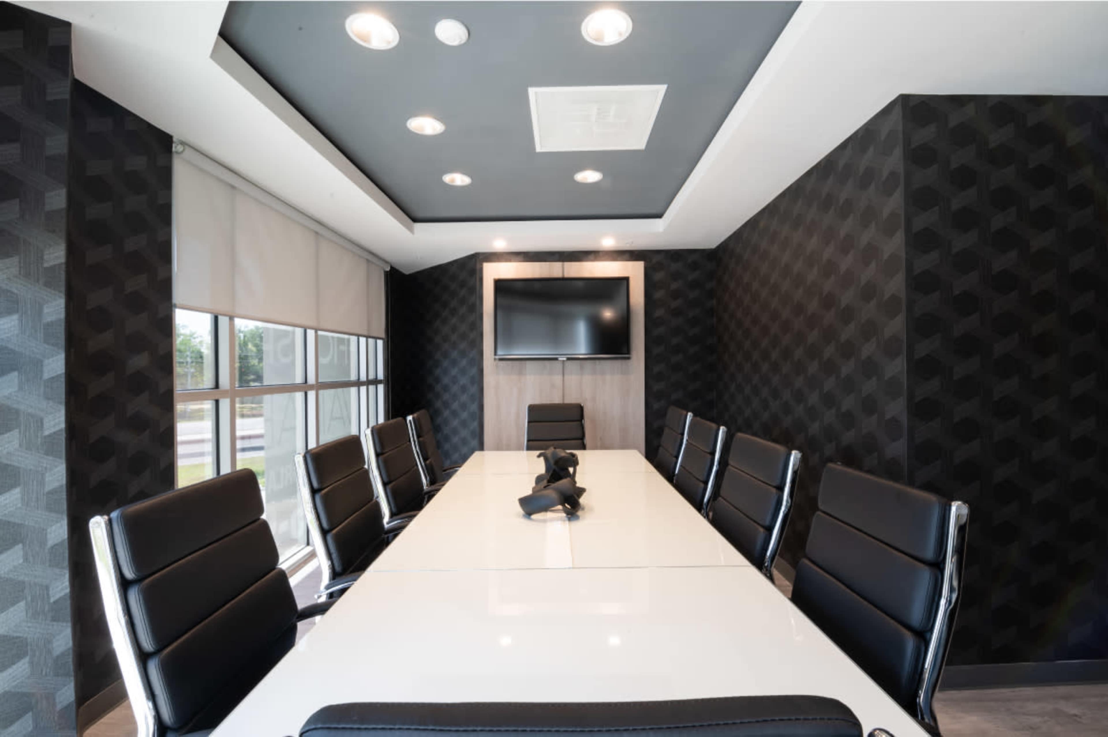 A modern conference room features a long white table surrounded by black chairs, with a wall-mounted television and geometric patterned wallpaper.