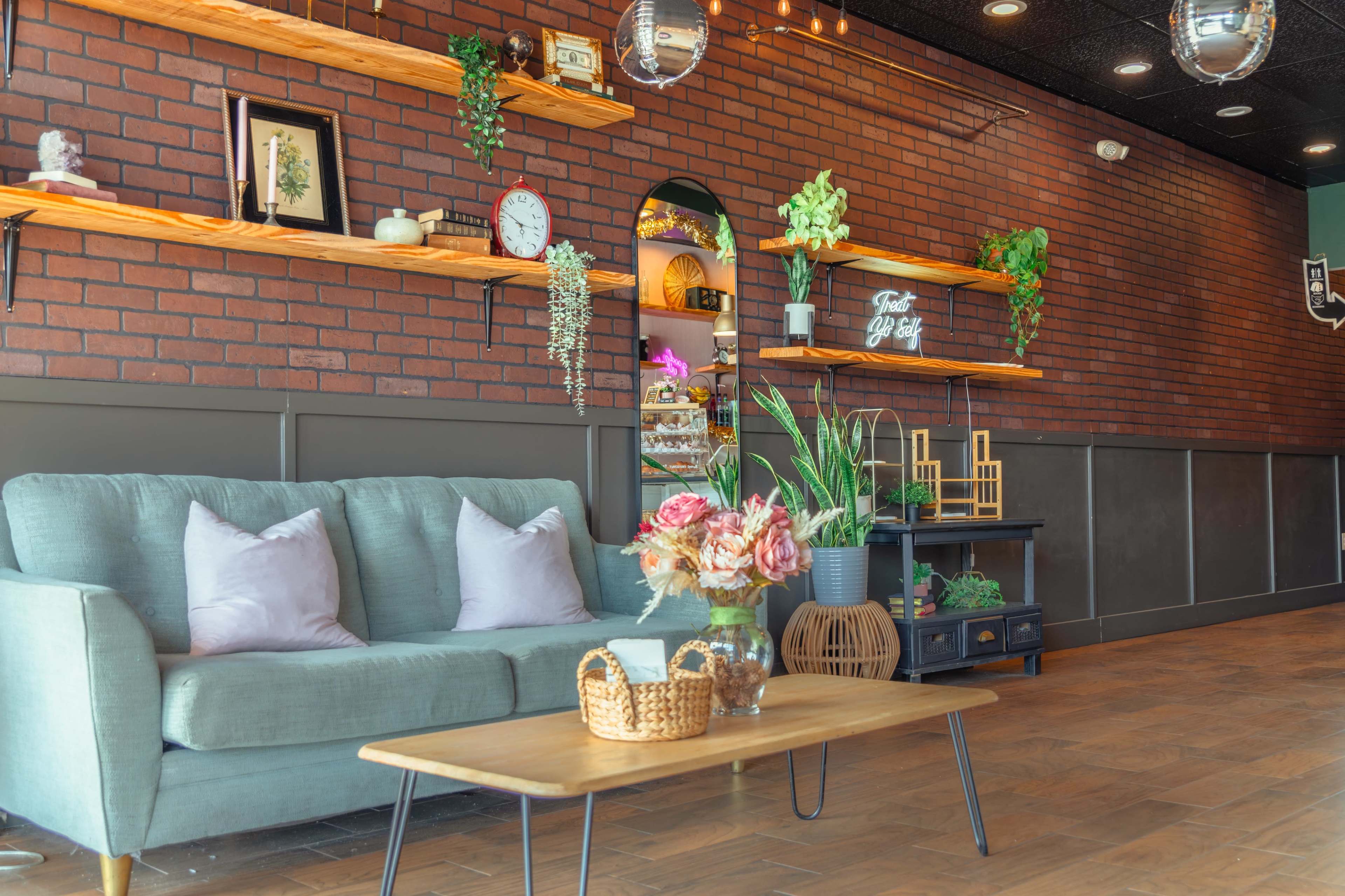 The image depicts a cozy café interior featuring a green sofa, a wooden coffee table, and a wall with brick accents adorned with plants and decorative shelves.