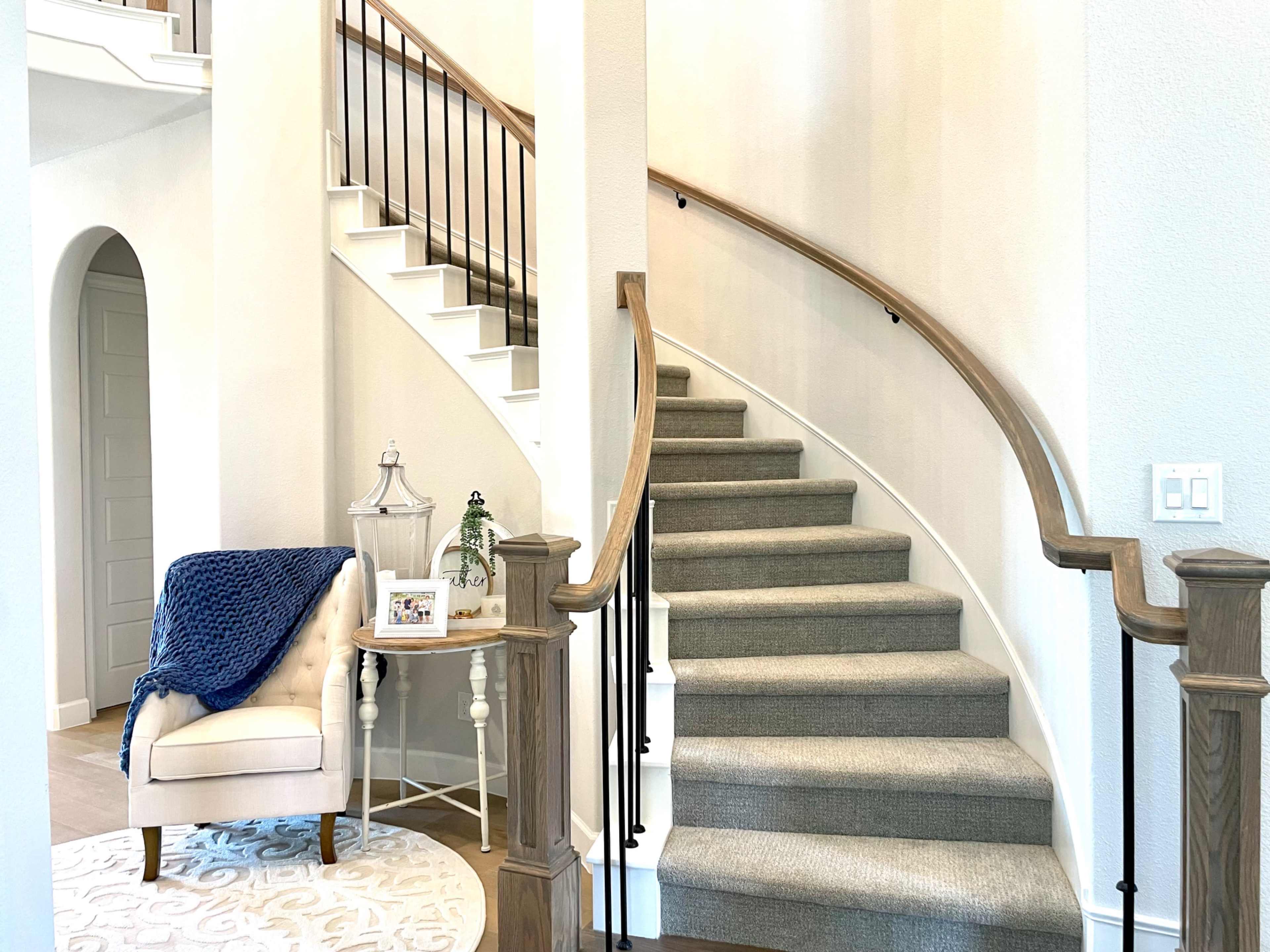A well-lit entryway featuring a curved staircase, a small armchair with a blue throw, and a round table with decorative items.
