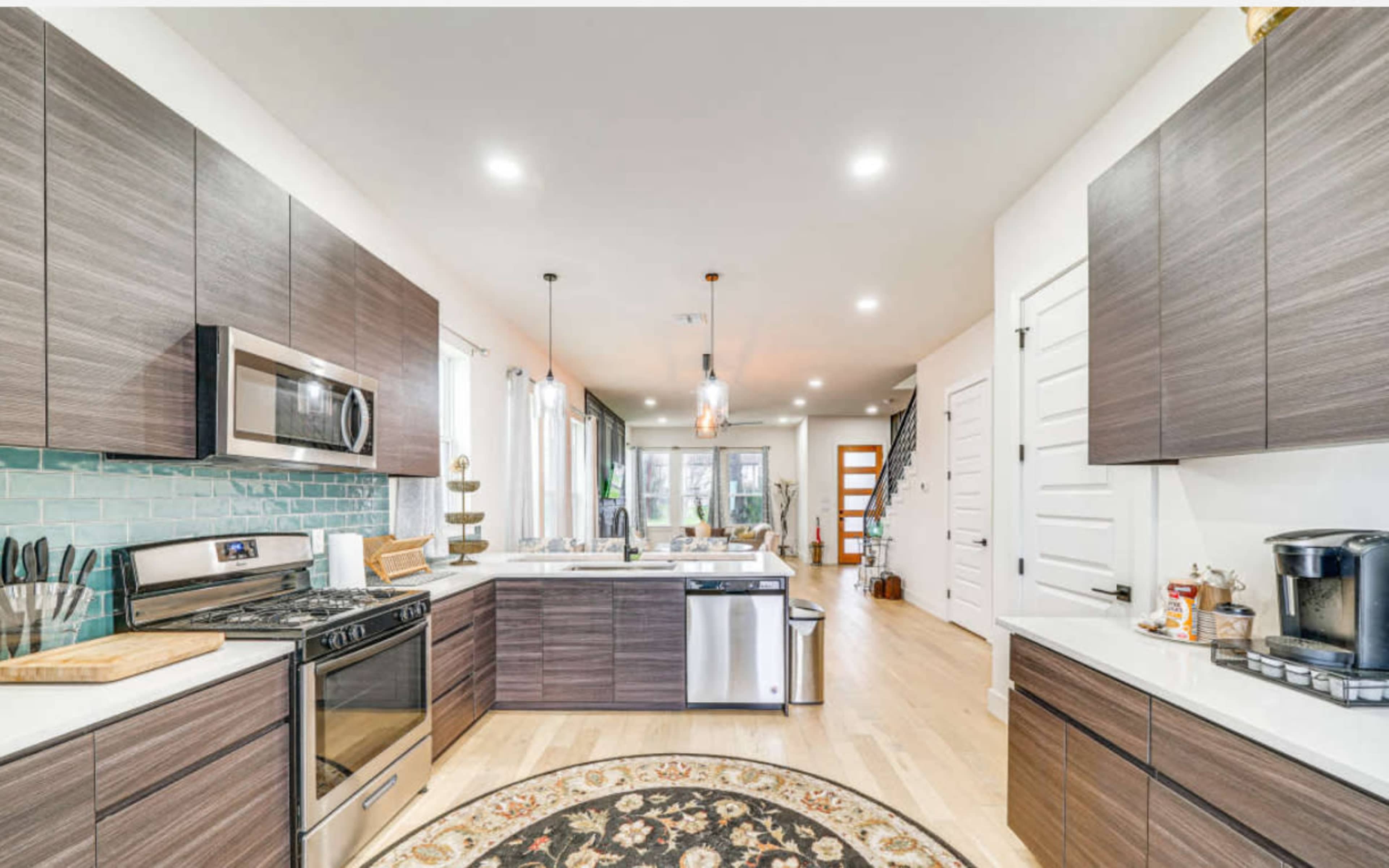 The image shows a modern kitchen with dark wood cabinets, stainless steel appliances, and a central island, alongside a living space with large windows and a staircase.