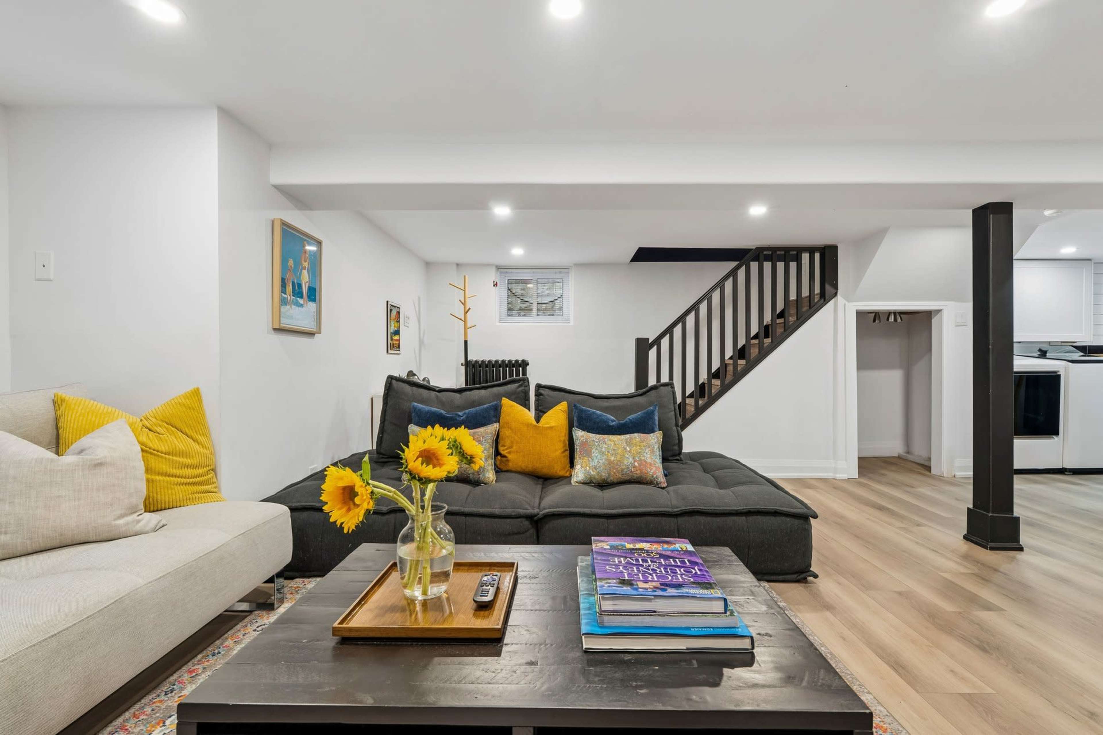 The image shows a modern living room with a dark sofa adorned with colorful pillows, a coffee table with books and a small vase of sunflowers, and a staircase in the background.