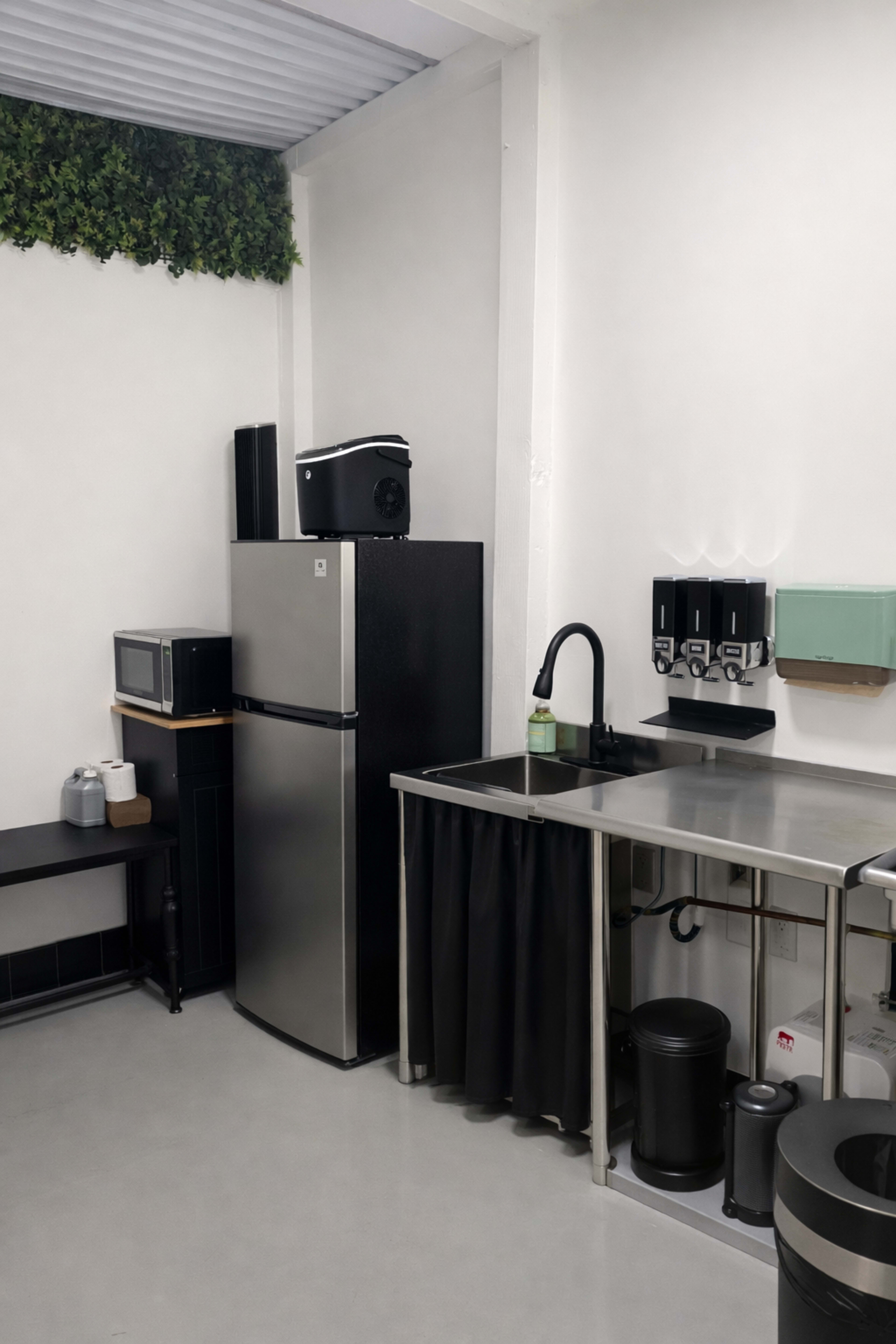 The image shows a modern kitchen with a stainless steel countertop, a black refrigerator, a microwave, and a series of black dispensers mounted on the wall.