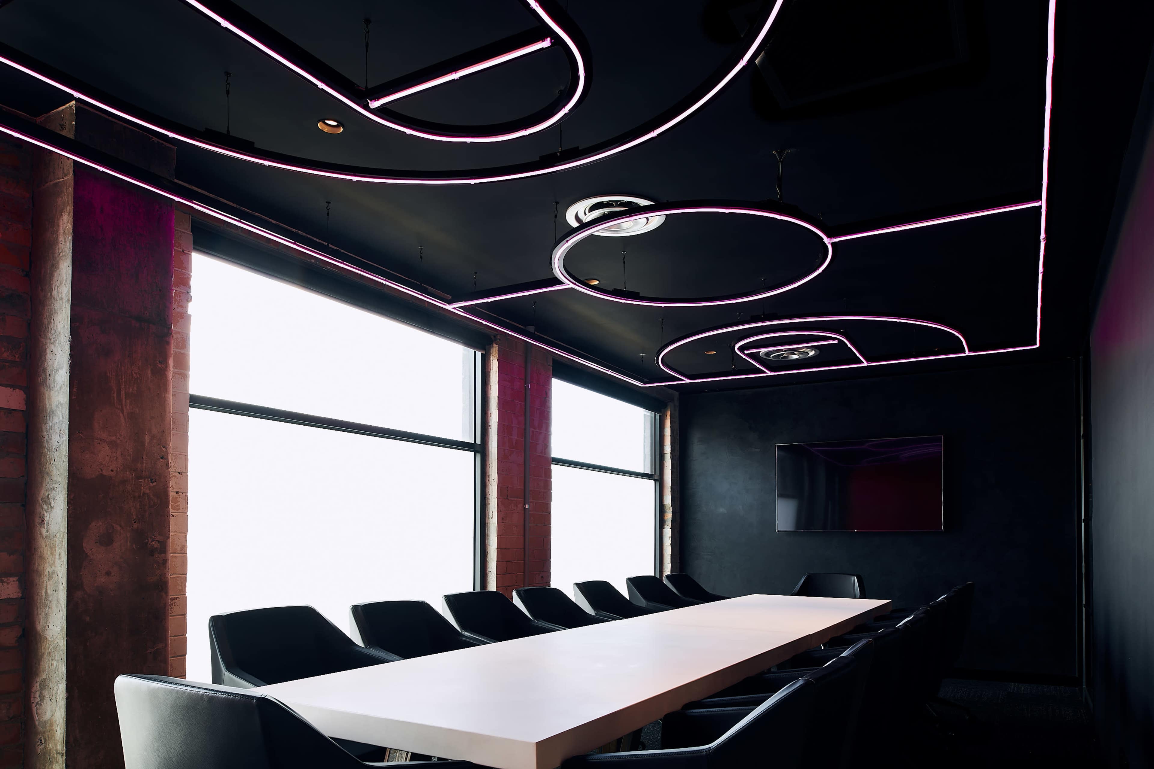 A modern conference room features a long white table, black chairs, and illuminated ceiling designs with pink neon lights.