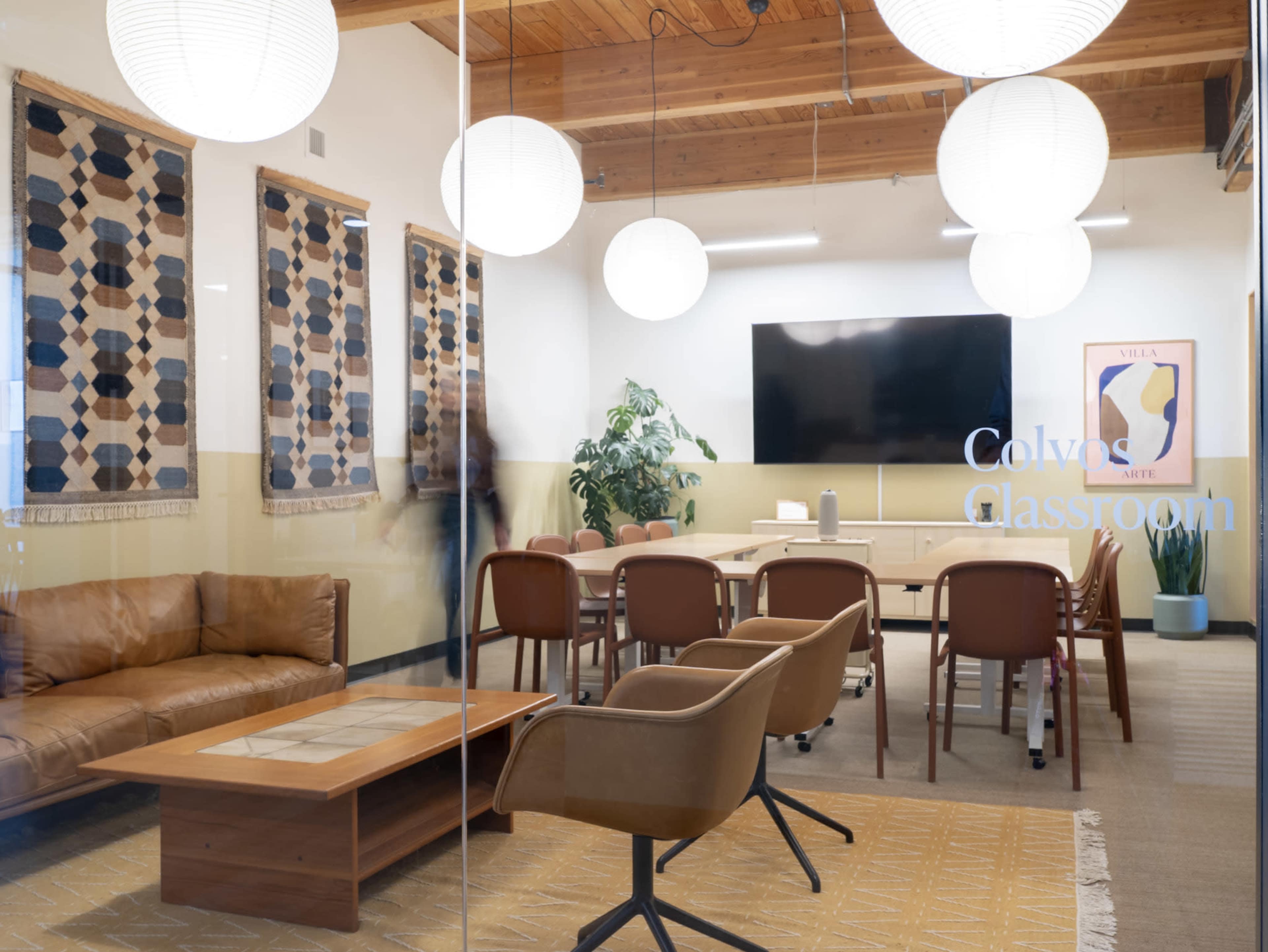 The image shows a modern classroom setting with a wooden ceiling, a large screen on the wall, a seating area with a brown couch, and several round light fixtures.