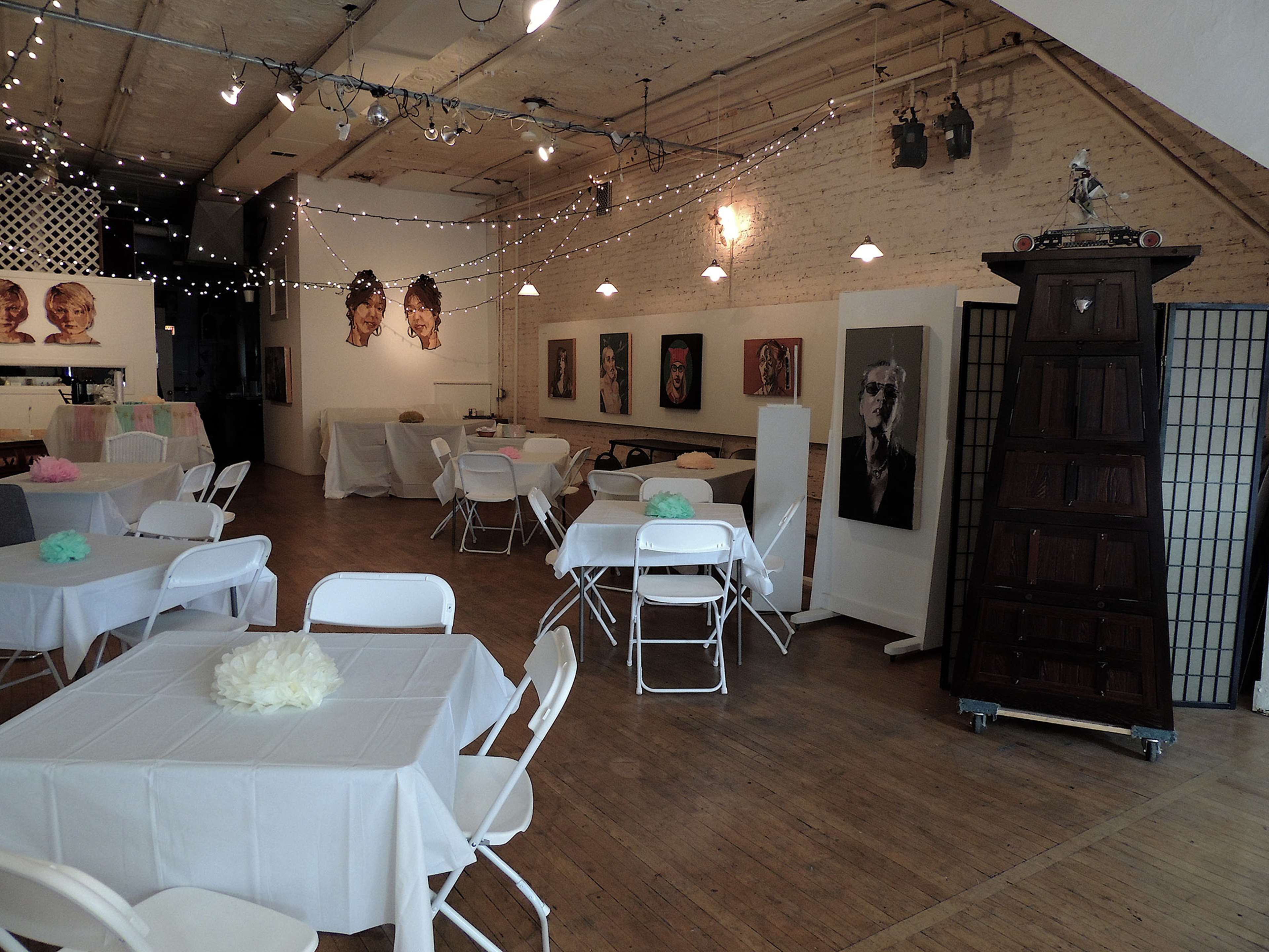 An event space with white tables and chairs, decorated with light-colored floral centerpieces, and featuring art on the walls and string lights overhead.