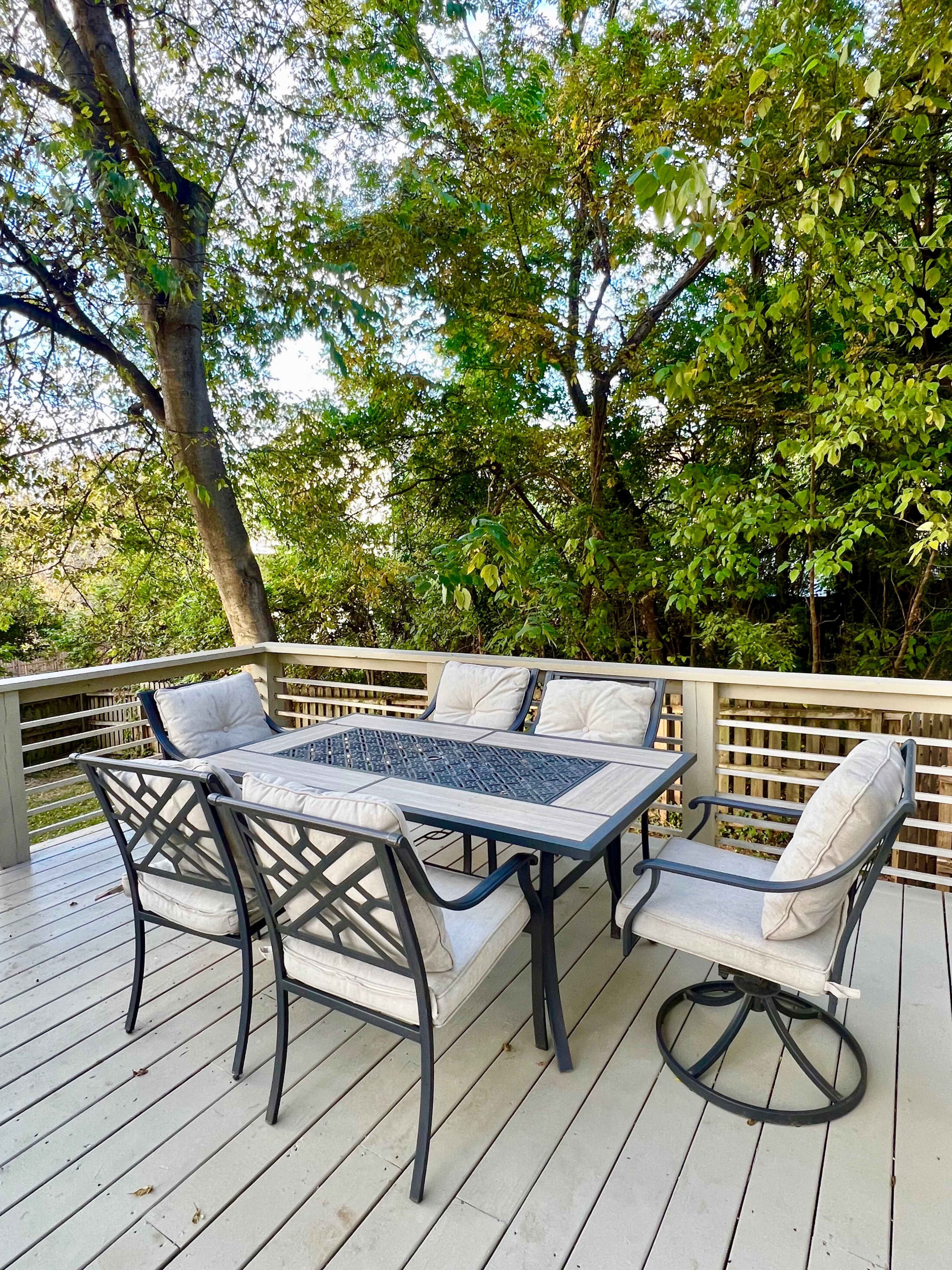 A patio dining set with a rectangular table is surrounded by four cushioned chairs on a wooden deck amid lush greenery.