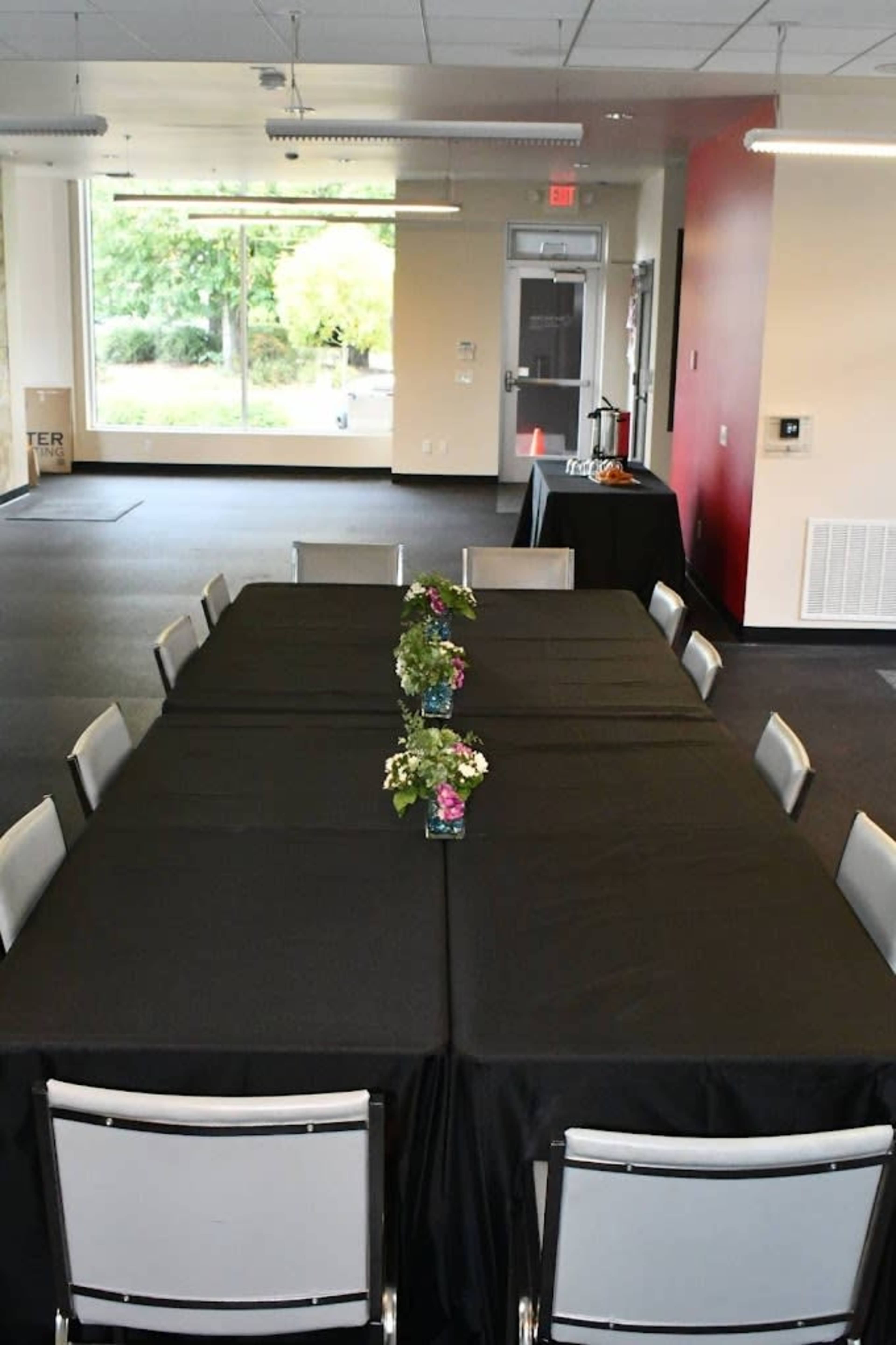 A long rectangular table covered with a black tablecloth is set up in an empty room with windows and potted plants at the center.
