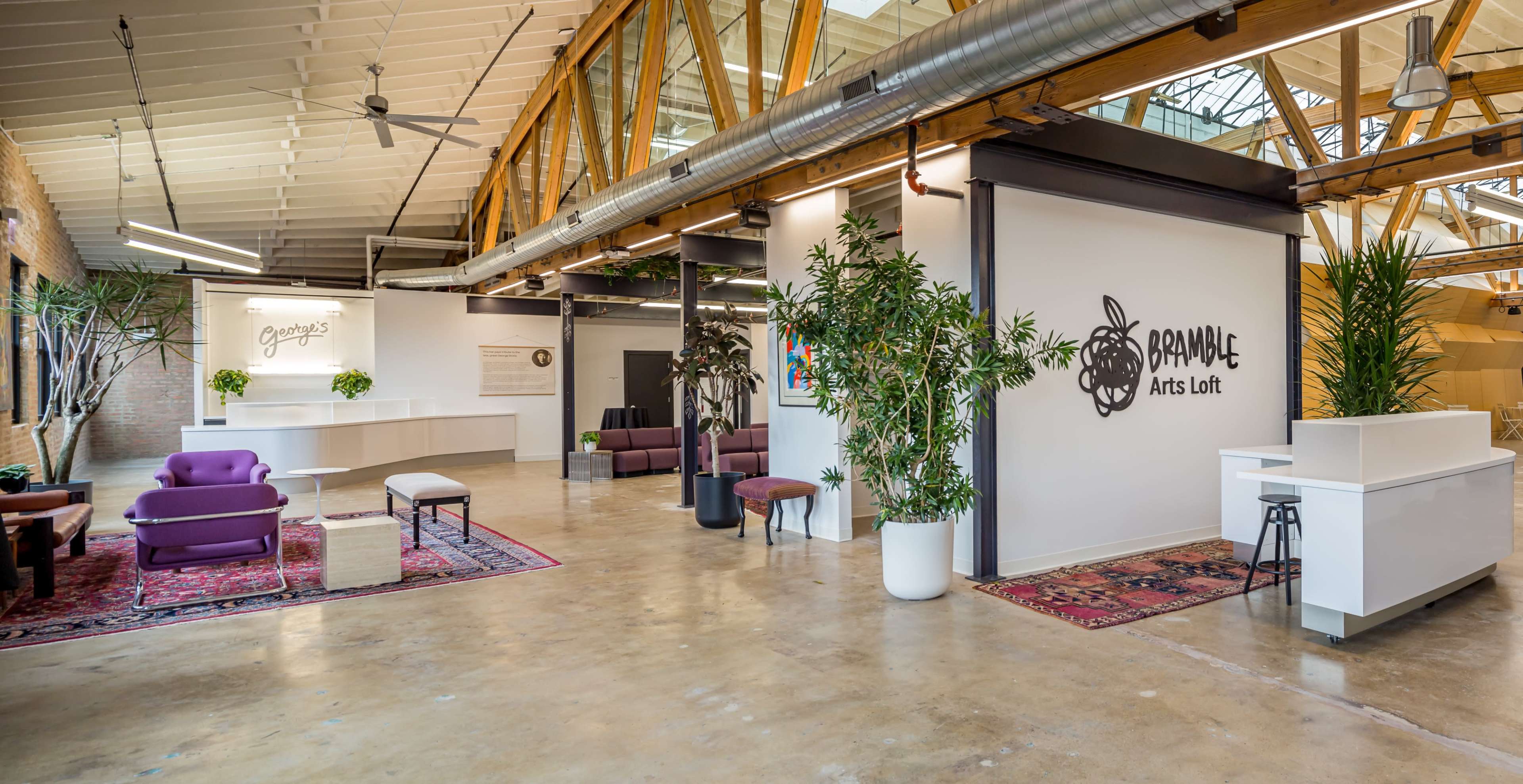 The image shows a modern, open interior space featuring a reception area, seating arrangements, and greenery, with exposed wooden beams and large windows.