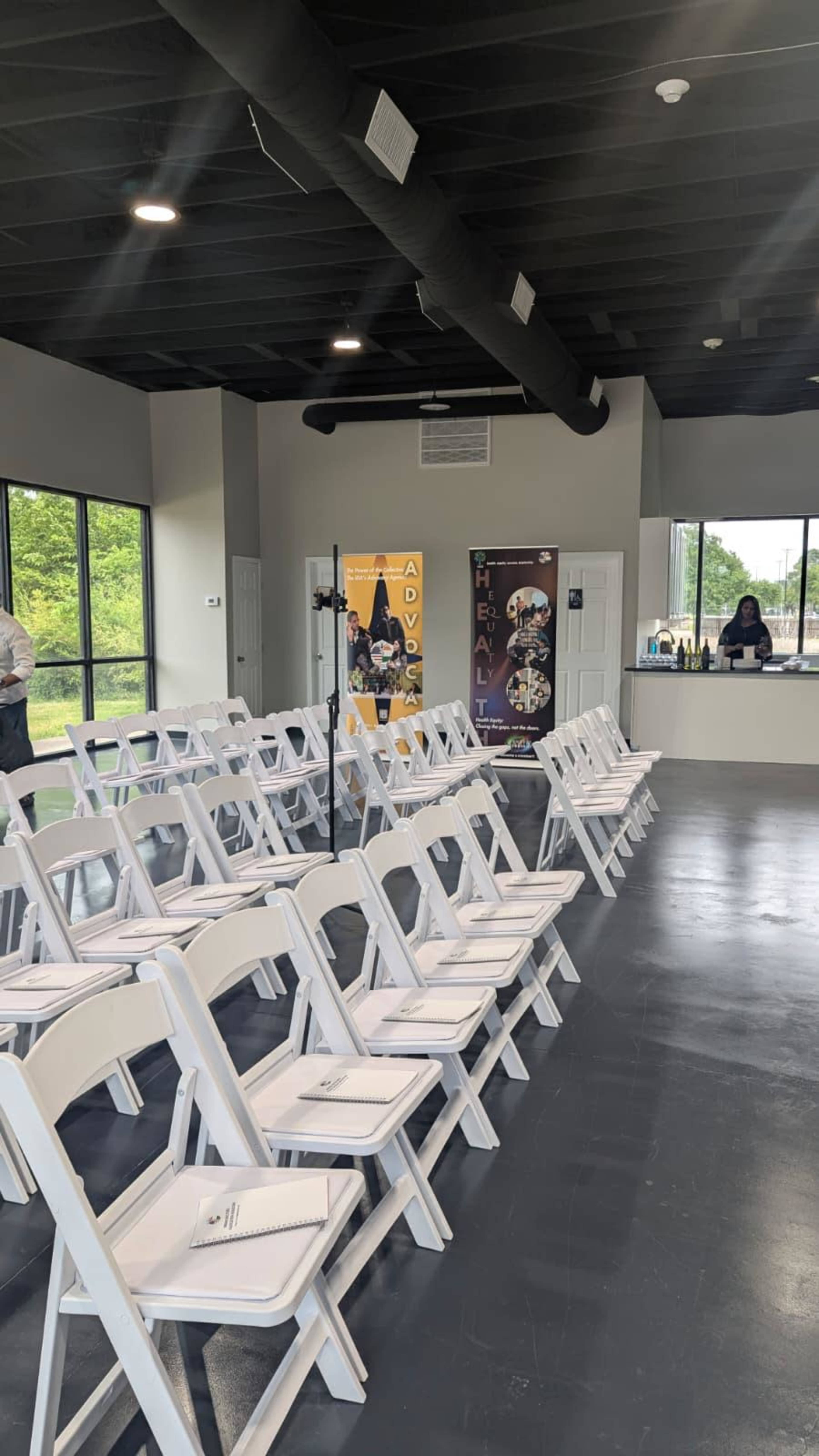 The image shows a spacious room set up for an event, featuring rows of white chairs facing a counter, with banners displayed on the walls.