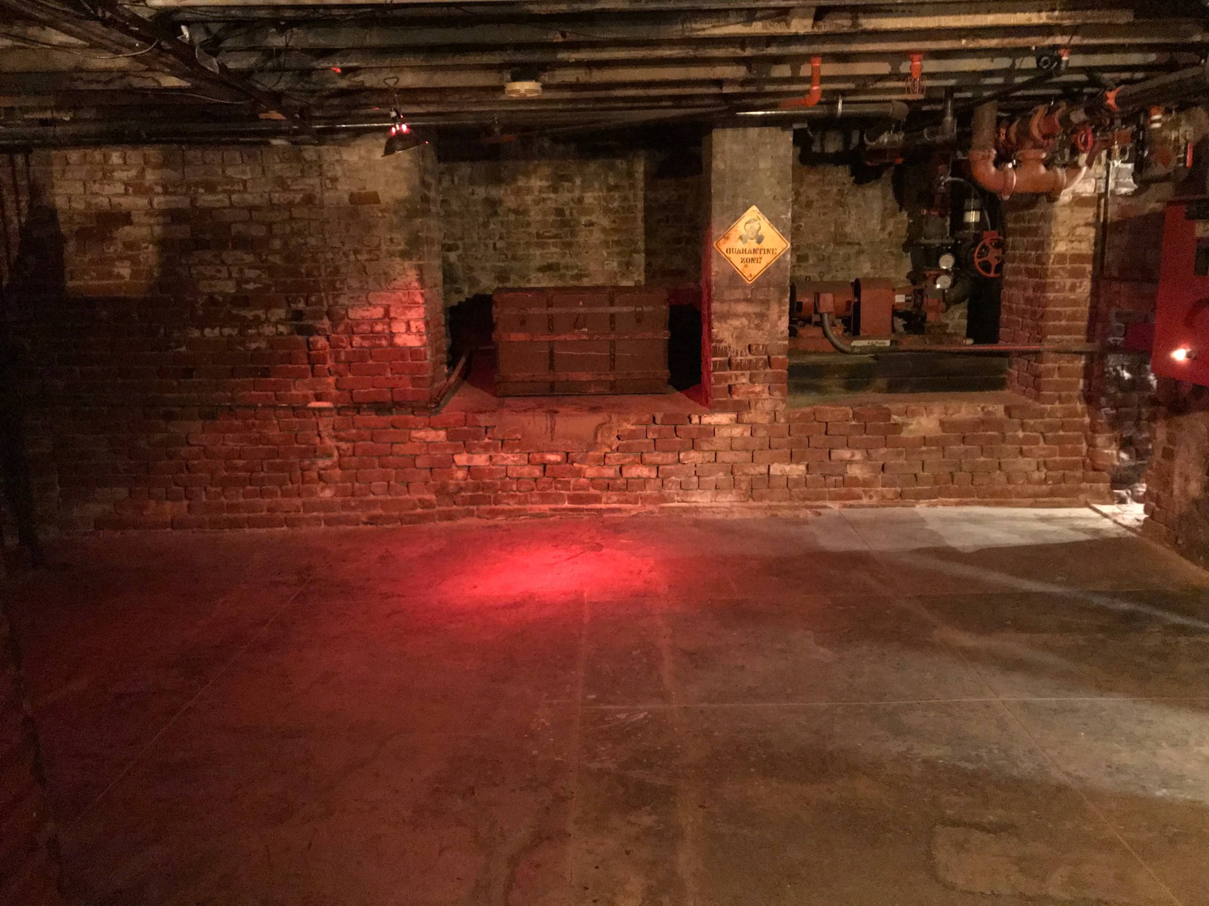 A dimly lit basement area with exposed brick walls, a concrete floor, and various industrial pipes and machinery in the background.