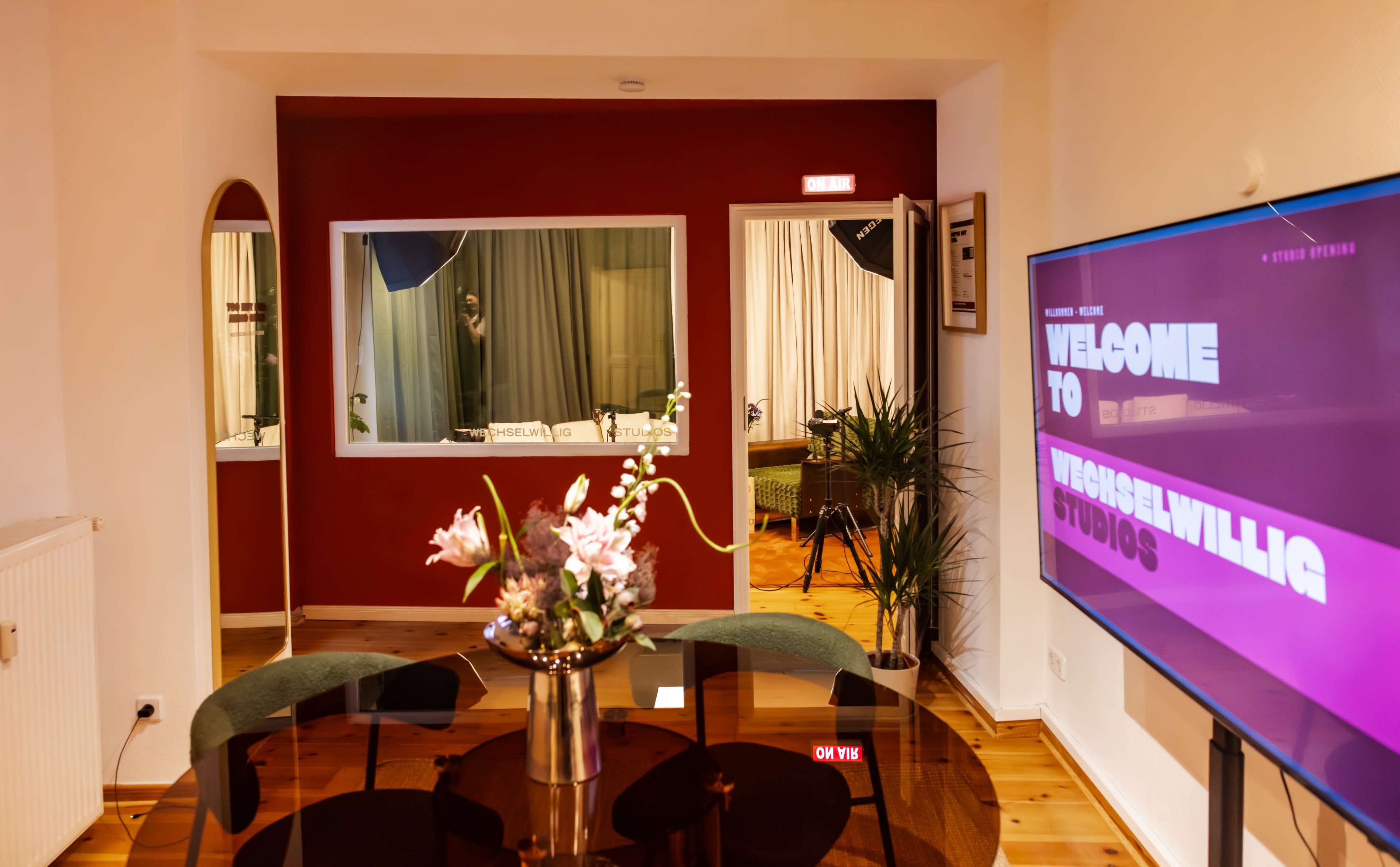 The image shows a modern studio space with a round table, flowers in a vase, and a large screen displaying "WELCOME TO VERSCHWILLIG STUDIOS."