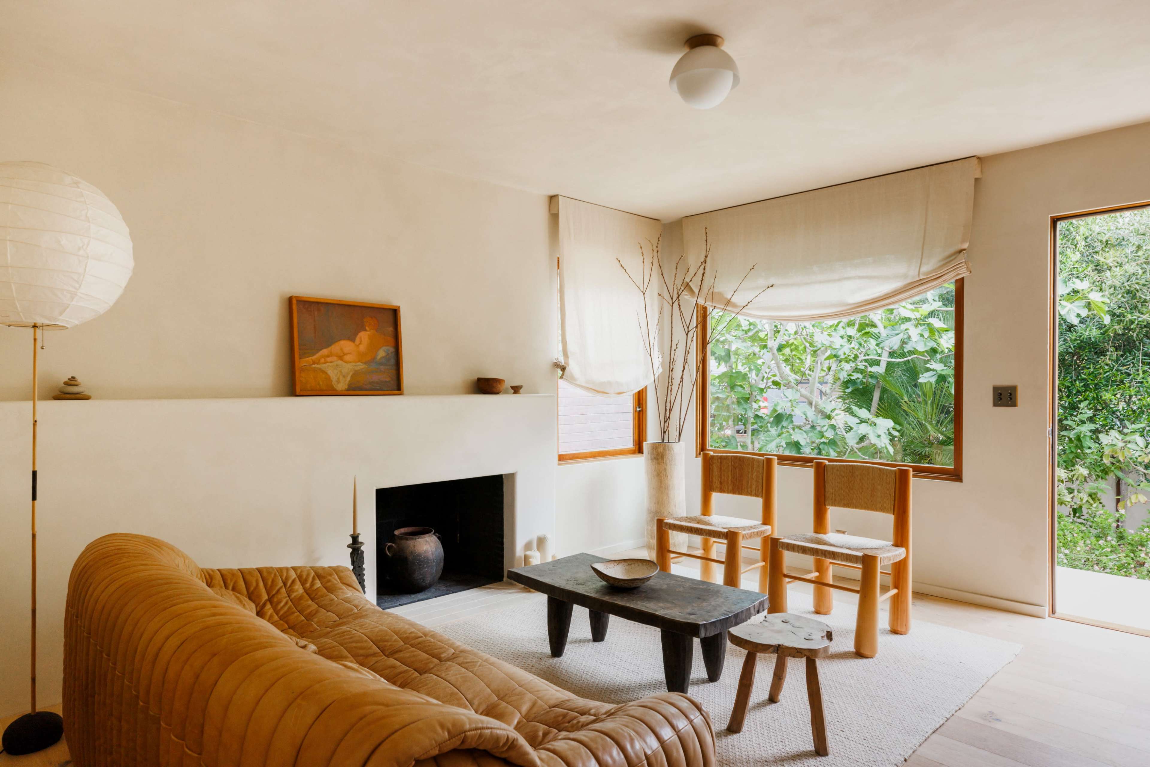The living room features a tan leather sofa, a wooden coffee table, two wooden chairs, and large windows letting in natural light with views of greenery outside.