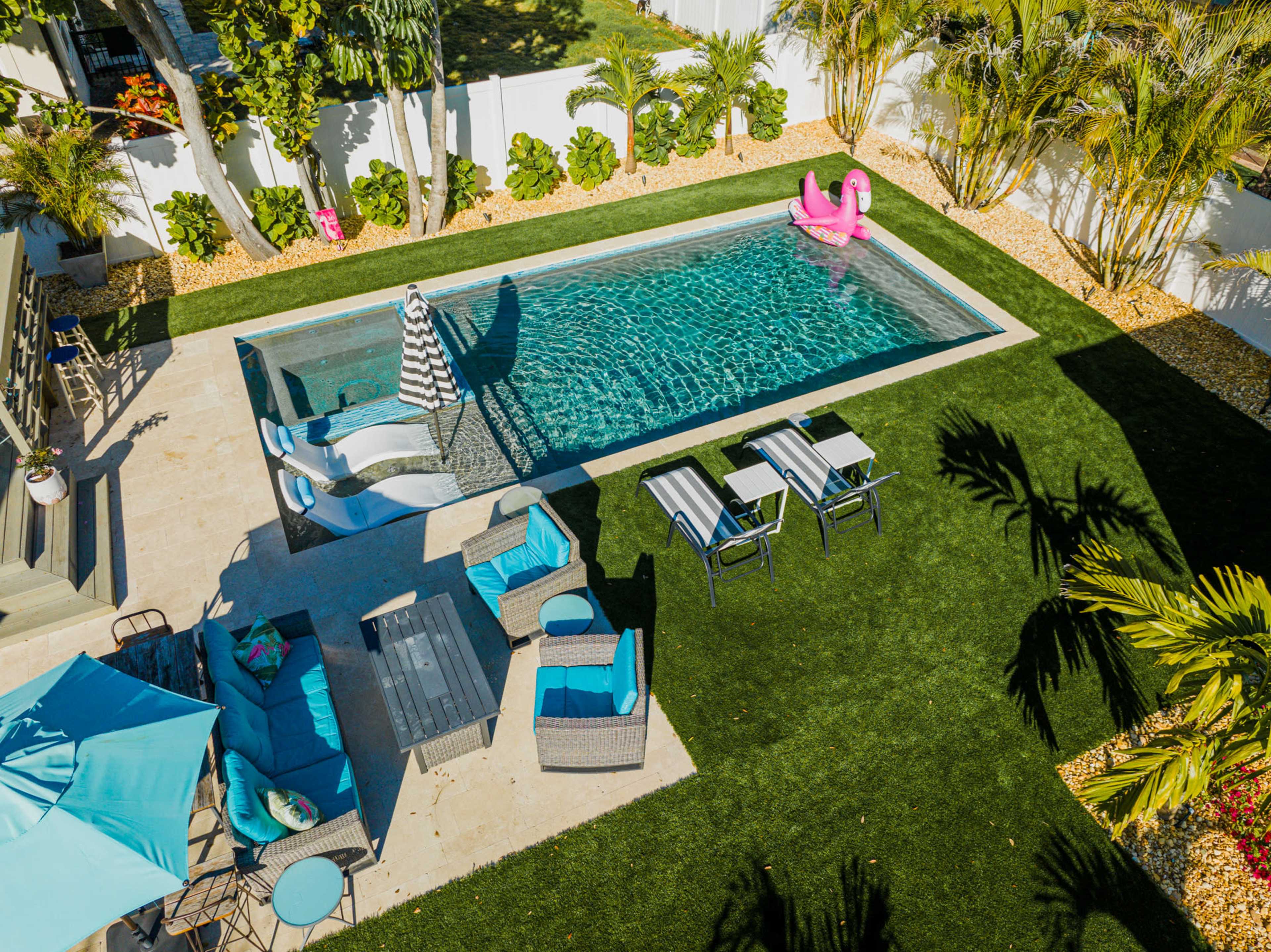 A modern backyard featuring a swimming pool surrounded by lounge chairs, a table set for outdoor dining, and tropical greenery.