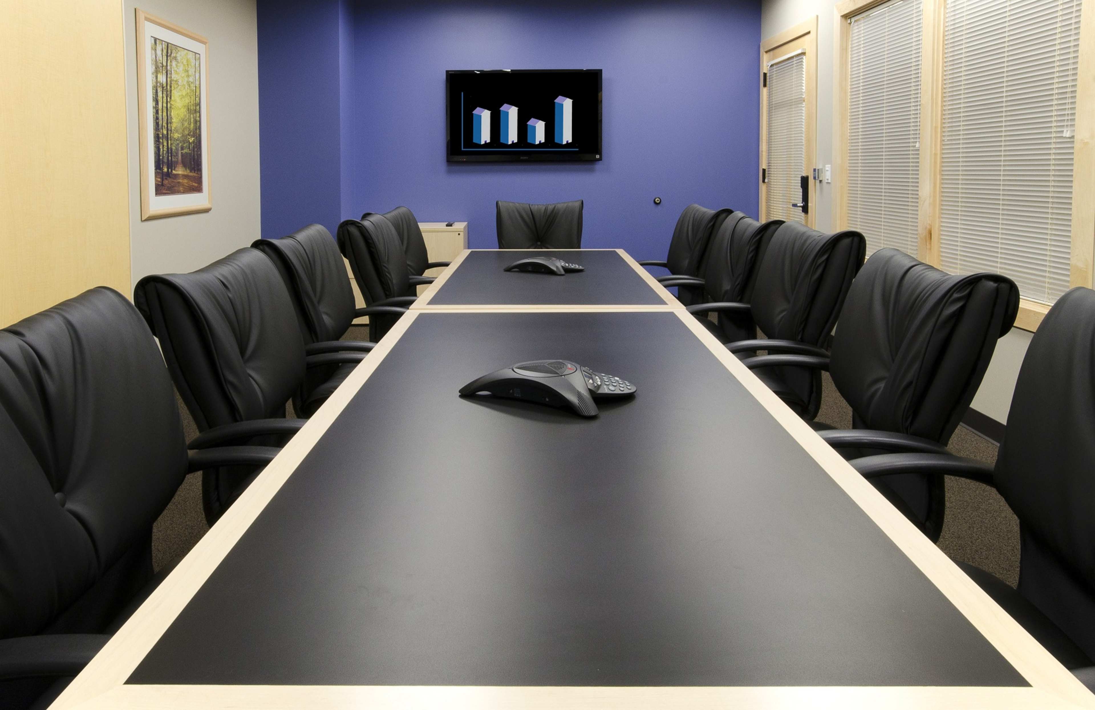 A conference room features a long black table with several black leather chairs, a video conference device in the center, and a wall-mounted screen displaying bar graphs.
