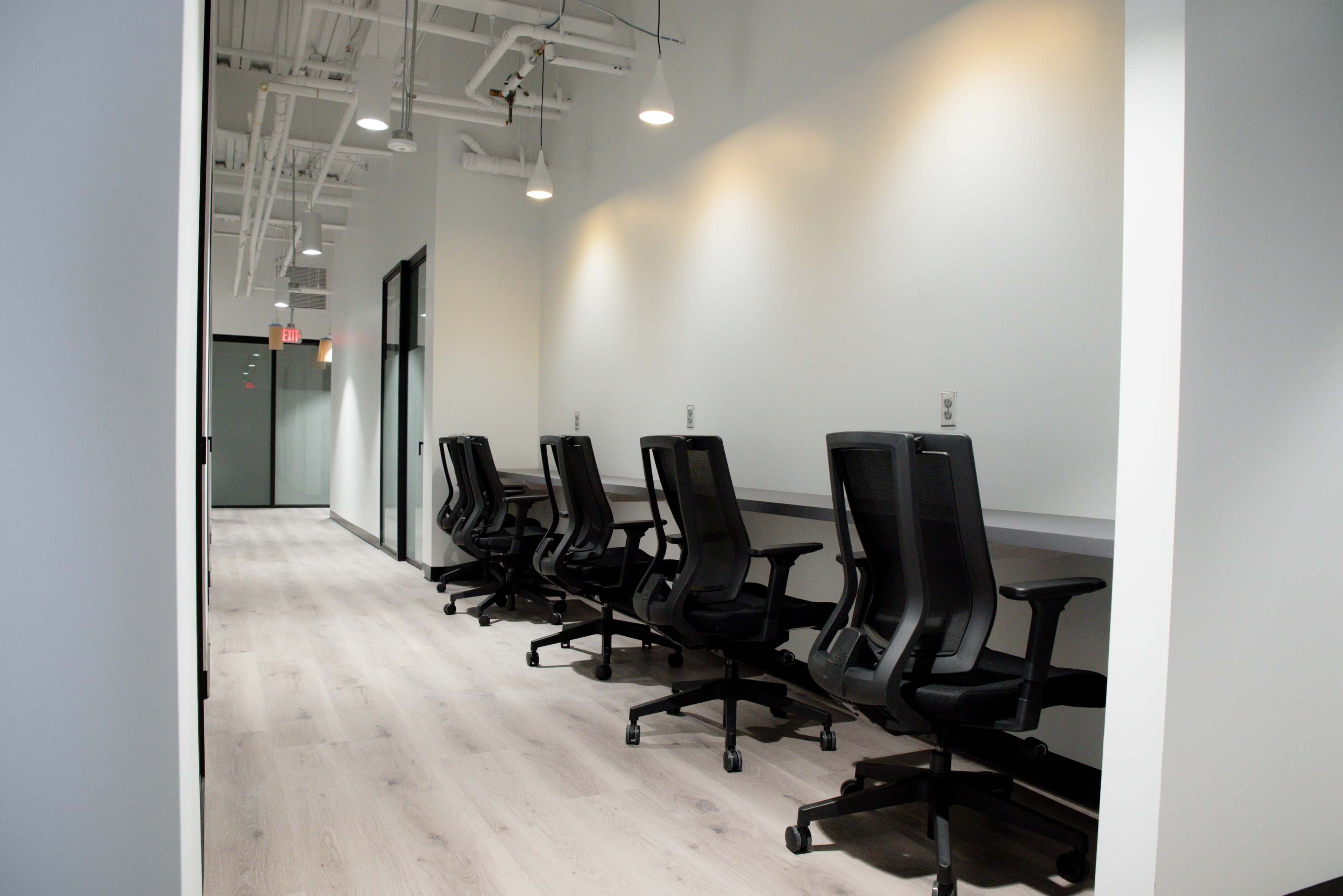 The image shows a row of empty black office chairs lined up against a light-colored wall in a modern workspace with wooden flooring.