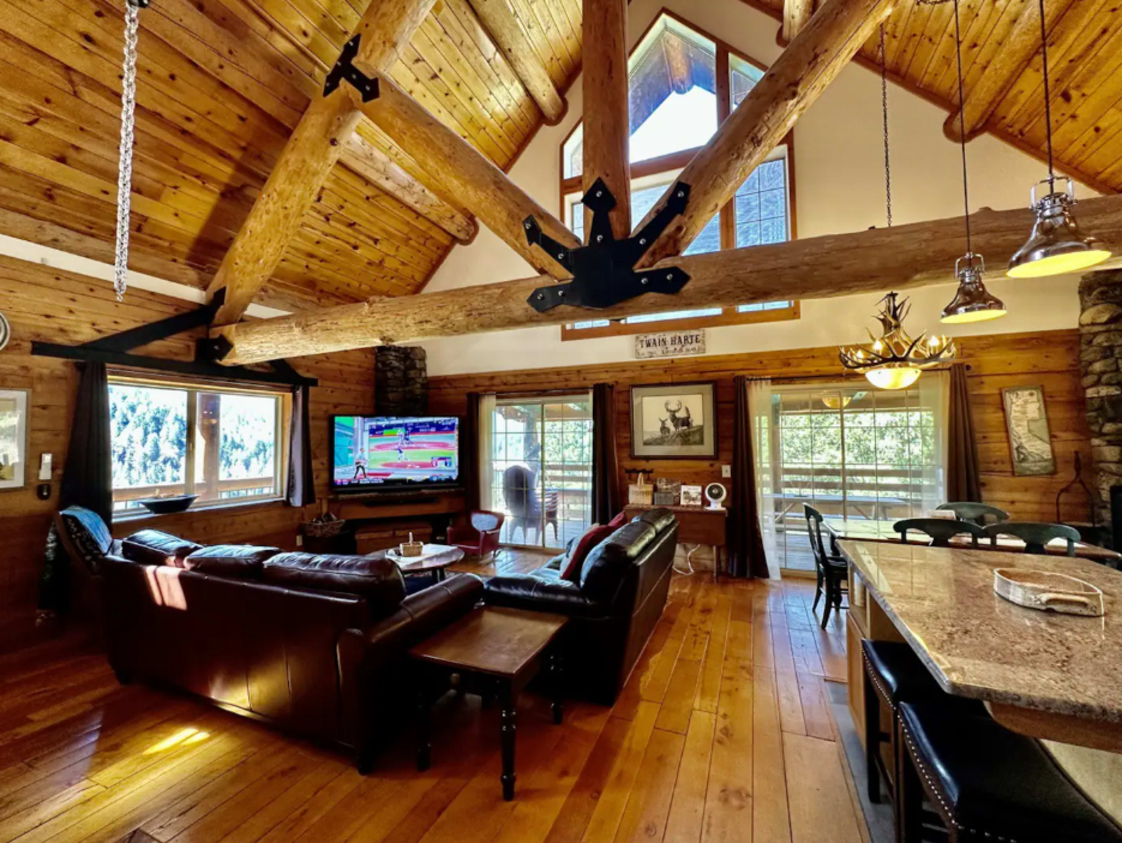 A spacious log cabin living room featuring a large leather sofa, a television, and large windows that offer views of the surrounding nature.