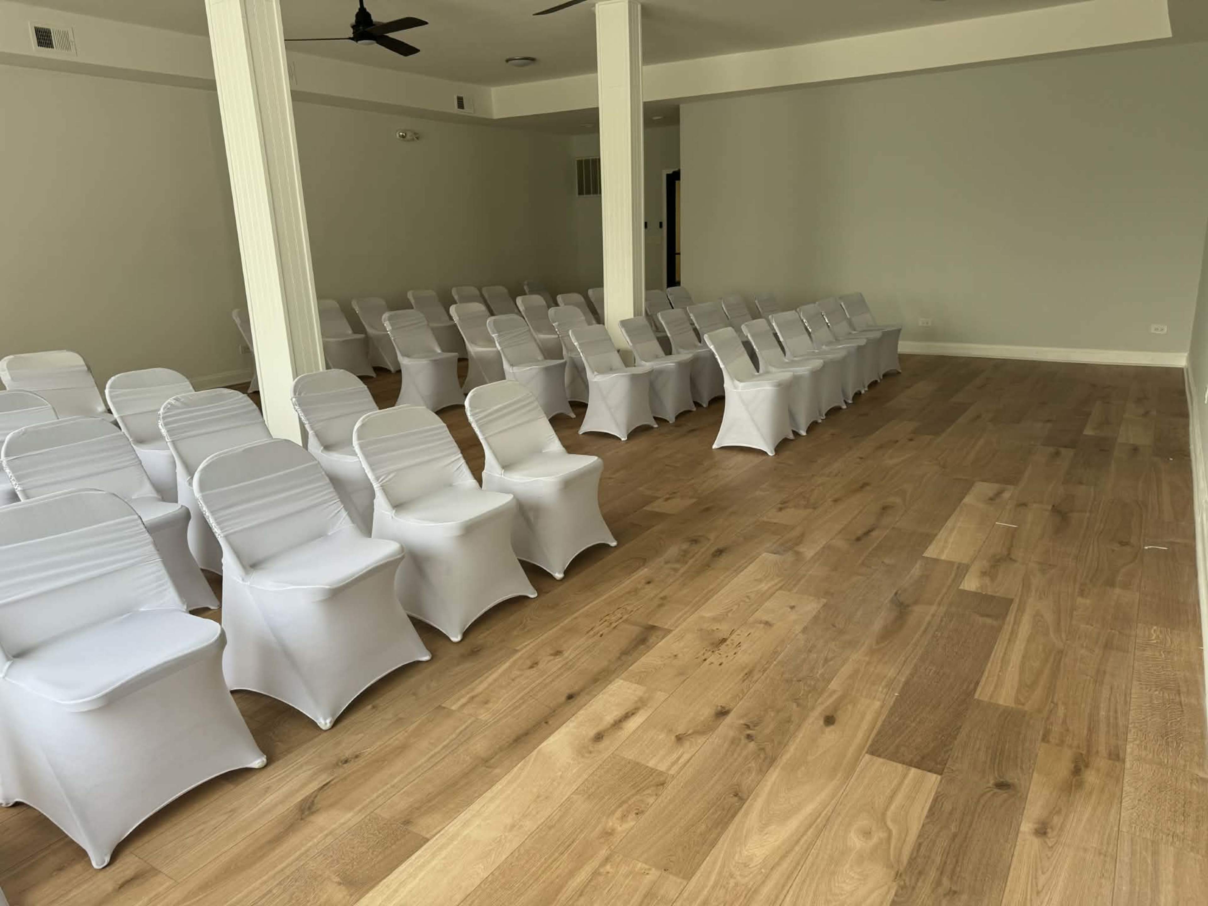 The image shows a spacious room with neatly arranged rows of white-covered chairs on a wooden floor.