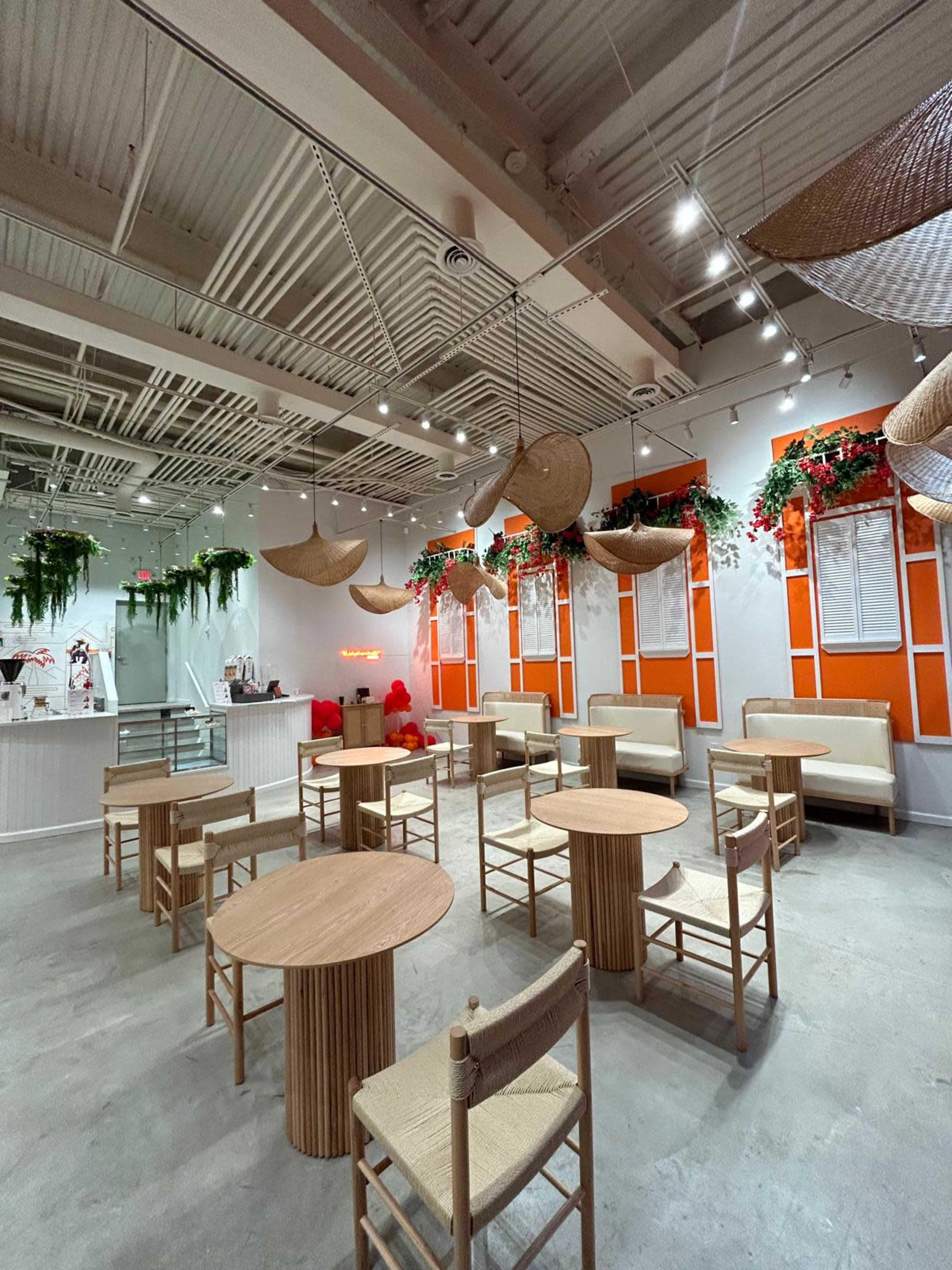 The image shows a modern café interior with wooden tables and chairs arranged around a spacious area, featuring vibrant orange accents and hanging decorative elements.