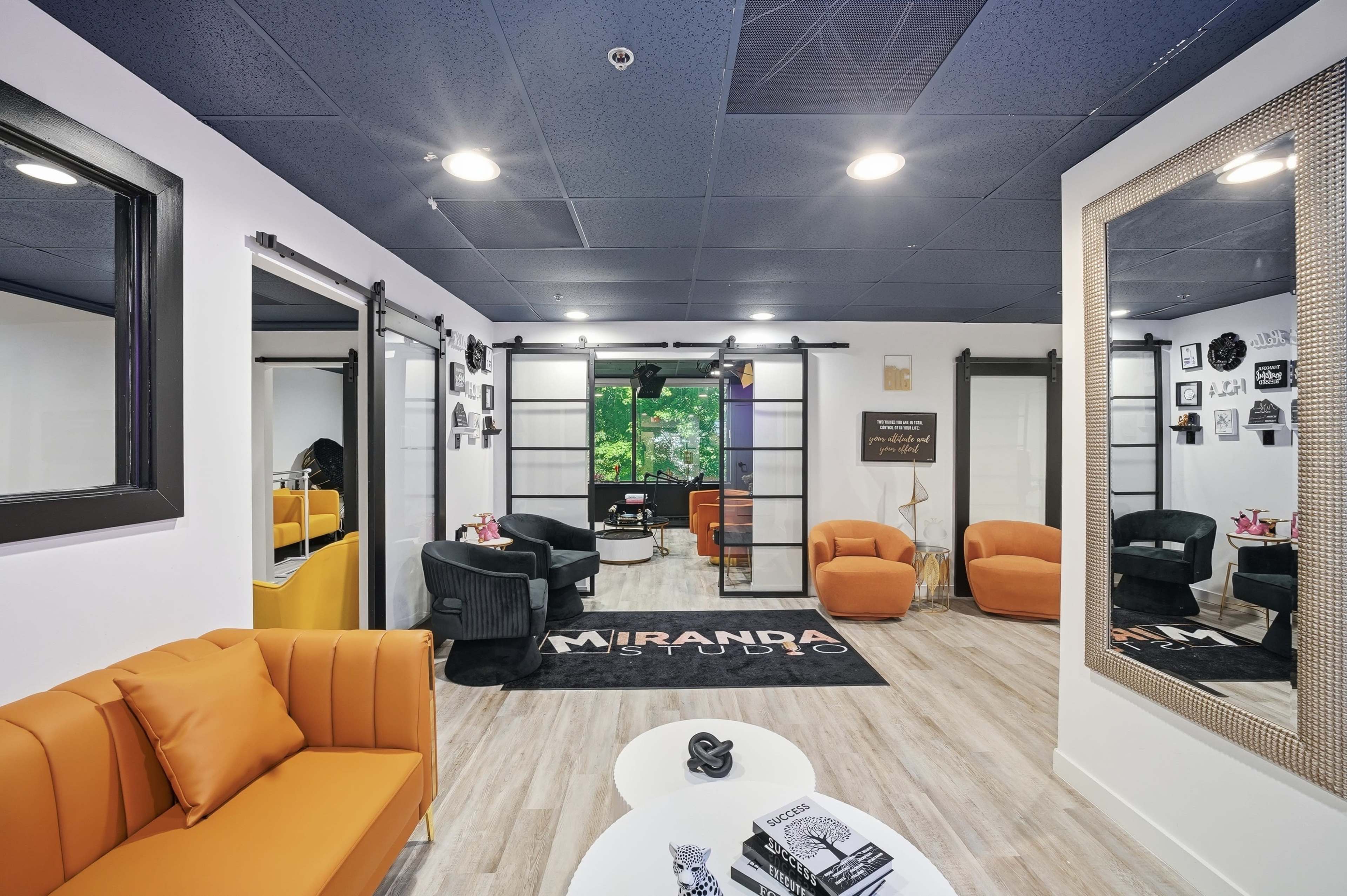 The image shows a modern studio interior featuring orange and black furniture, large mirrors, and a view of greenery outside through a window.