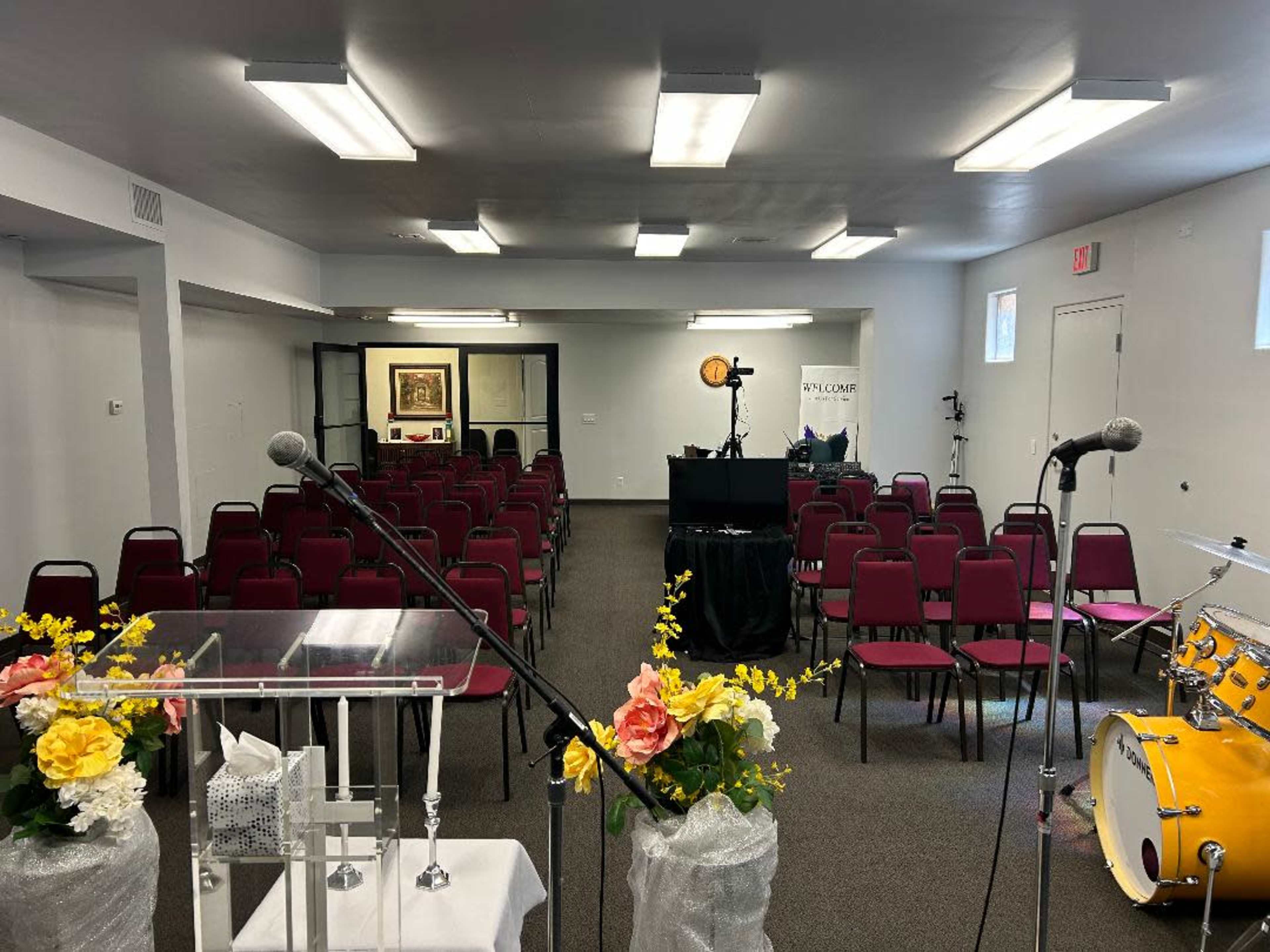 A spacious room set up for a gathering, with rows of red chairs facing a clear podium and a drum set arranged to the side.