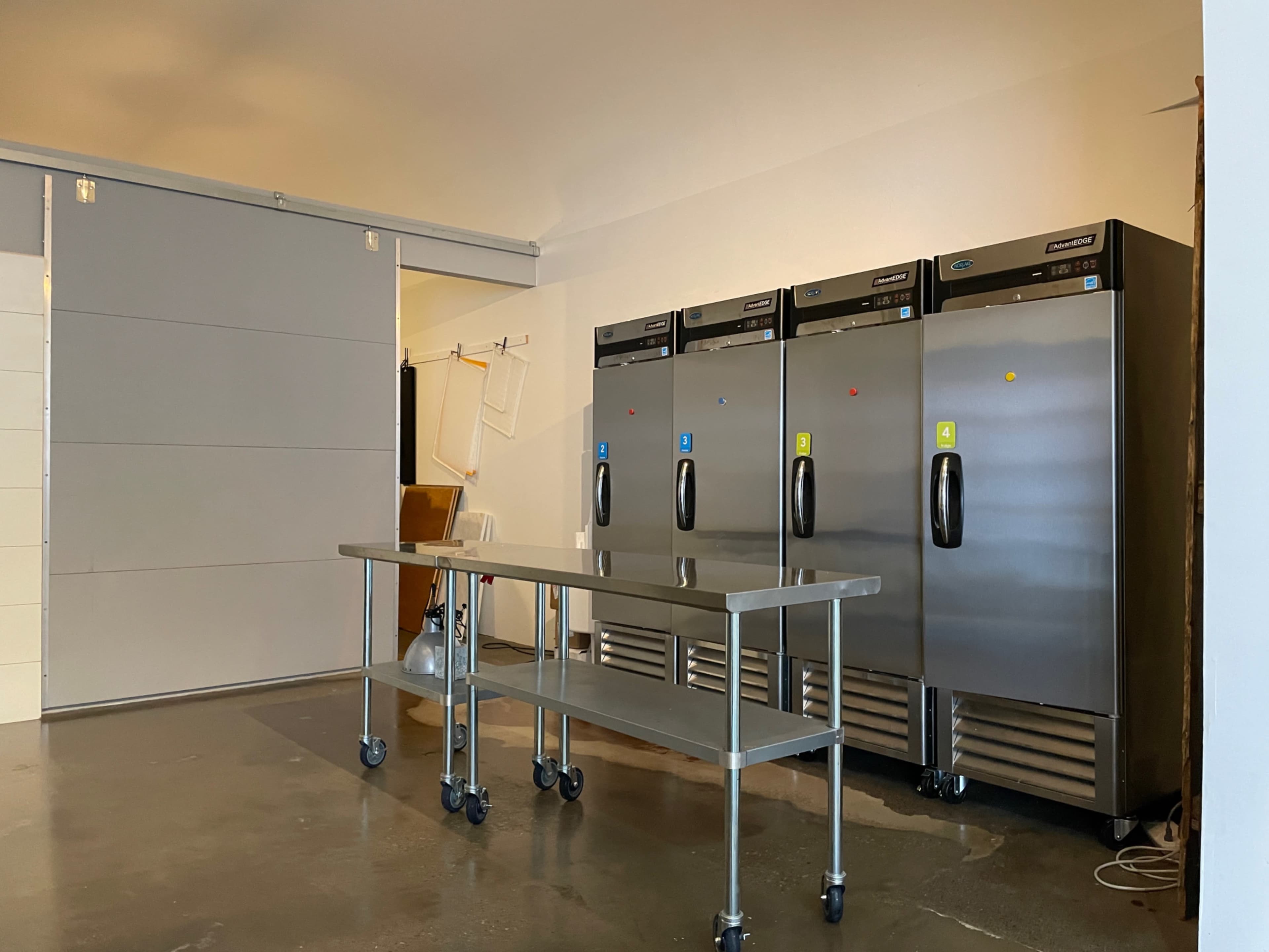 The image shows a commercial kitchen area with three stainless steel refrigerators and a stainless steel prep table on wheels.