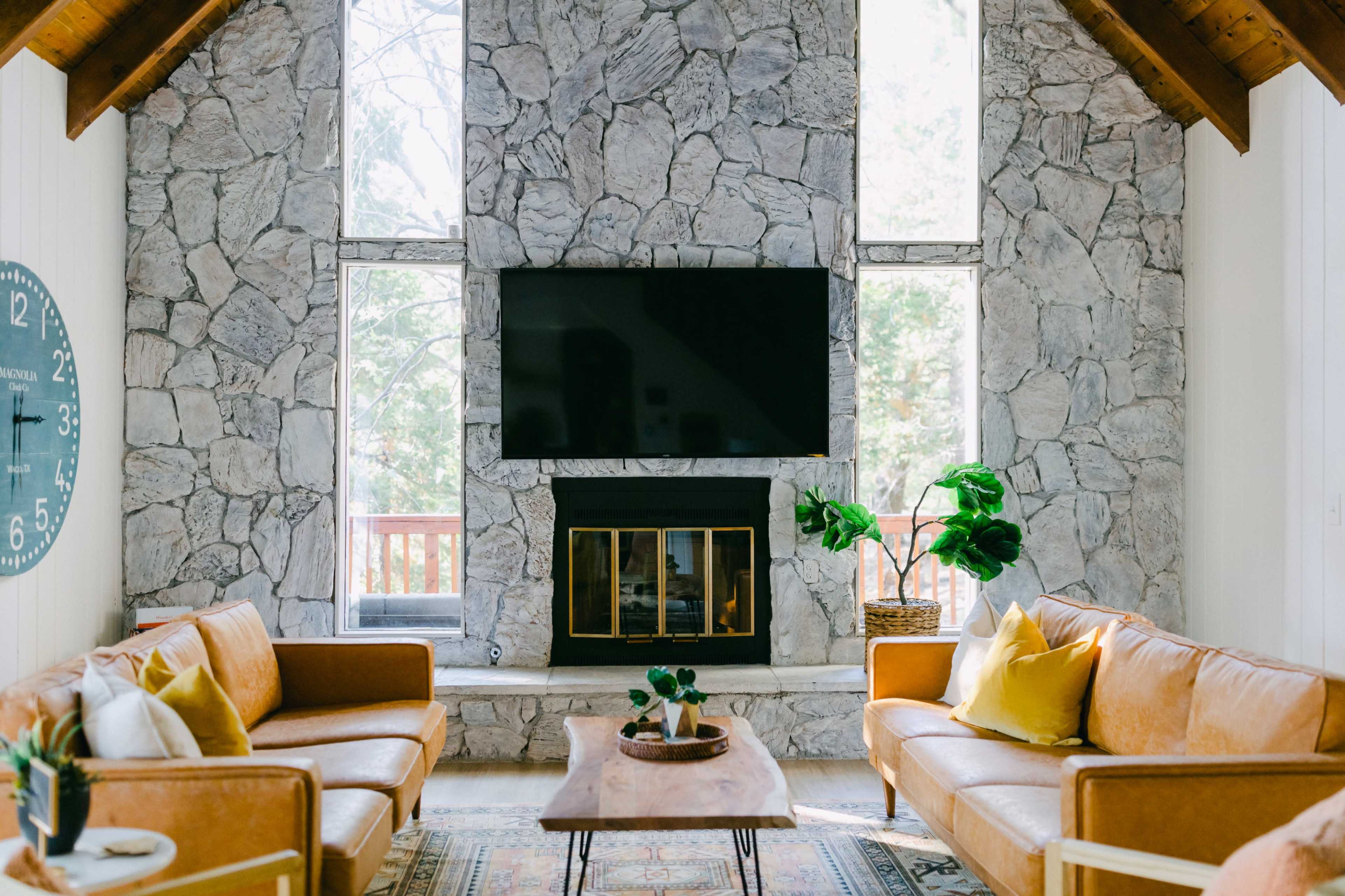 The image shows a living room featuring stone walls, a large flat-screen TV above a fireplace, and two leather sofas arranged around a wooden coffee table.