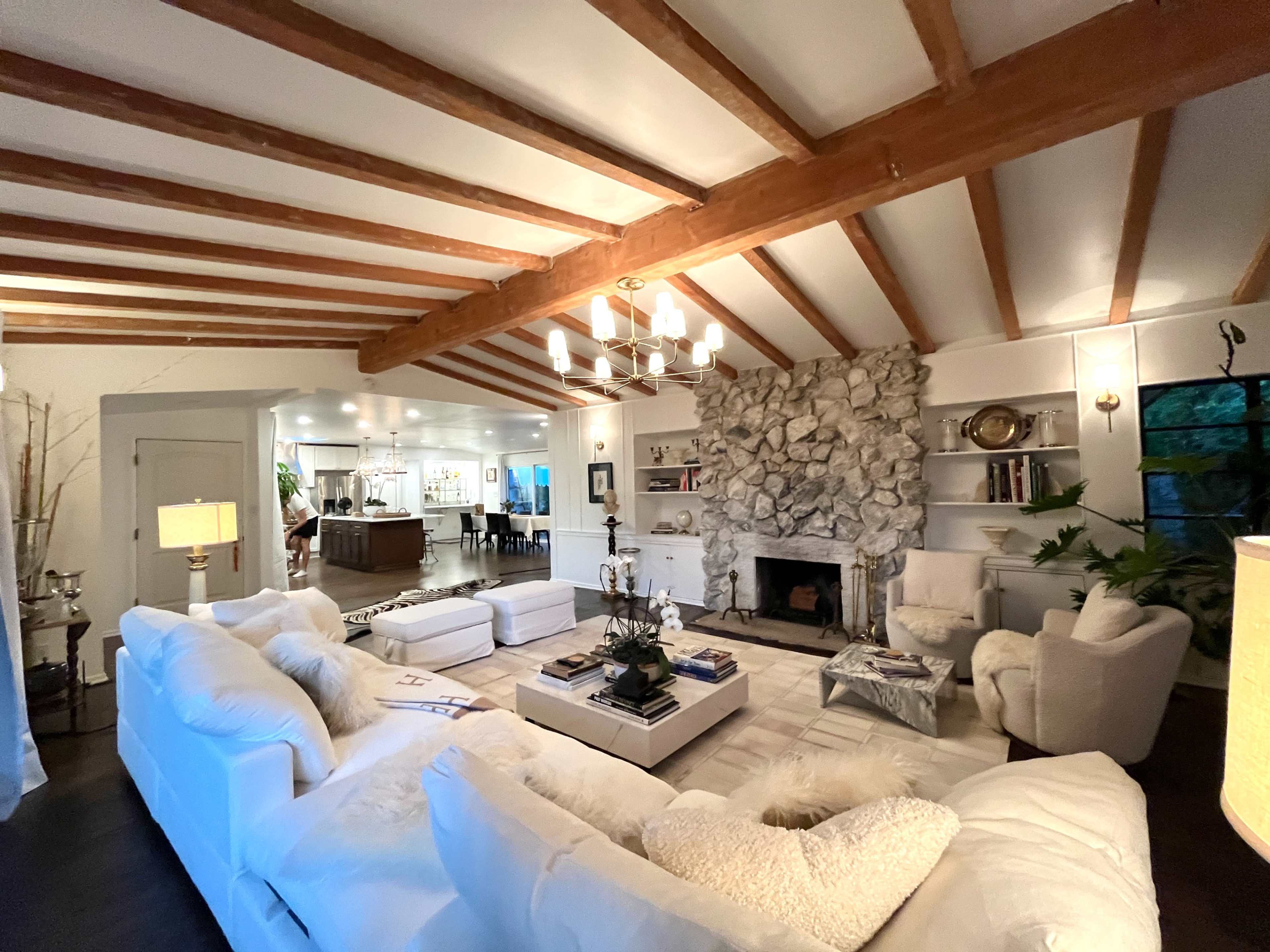 The image shows a spacious living room featuring a stone fireplace, white furniture, and exposed wooden beams on the ceiling.