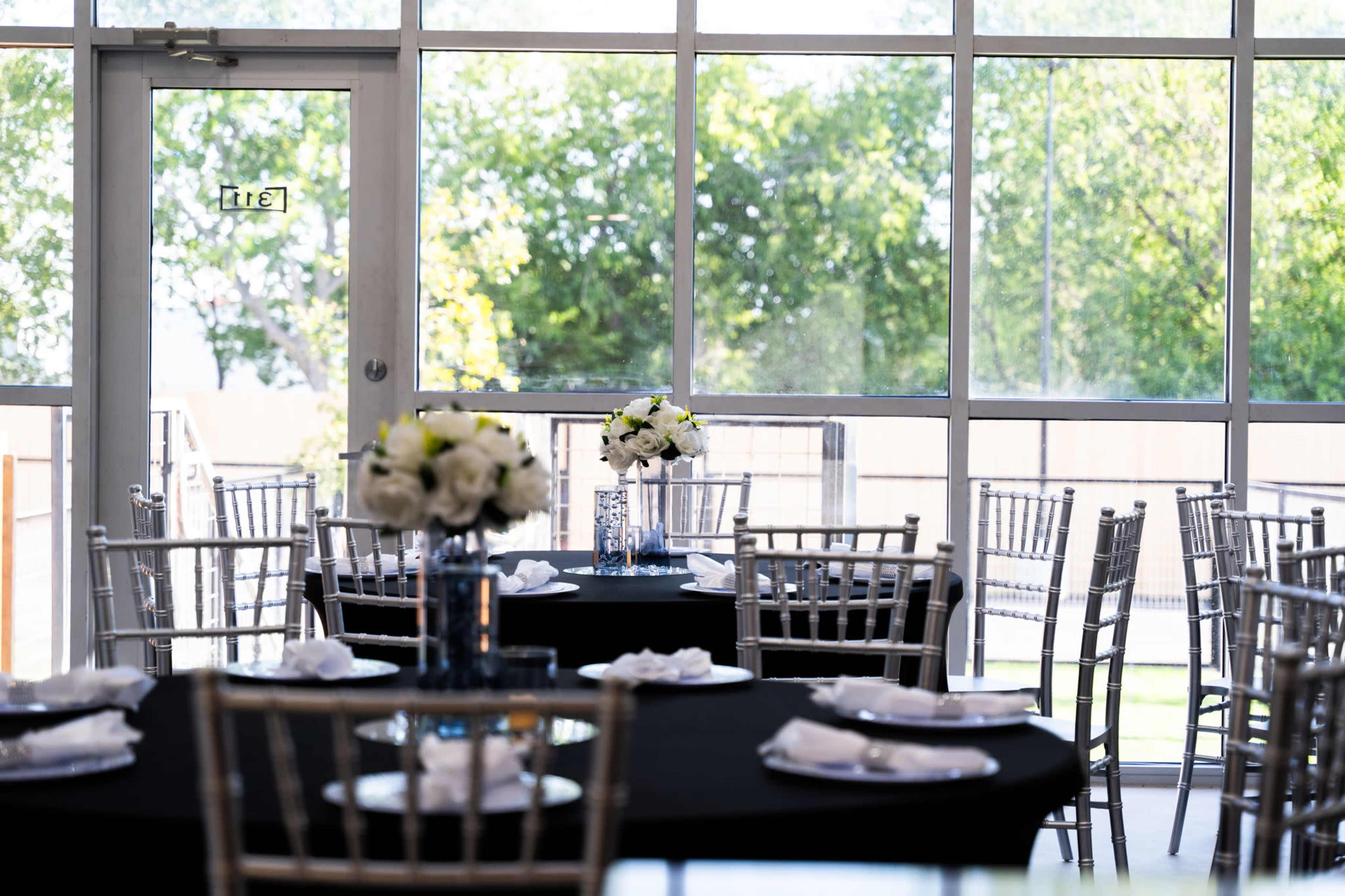 A large event space is set with round black tables adorned with white napkins and centerpieces of flowers, all surrounded by tall windows overlooking greenery.