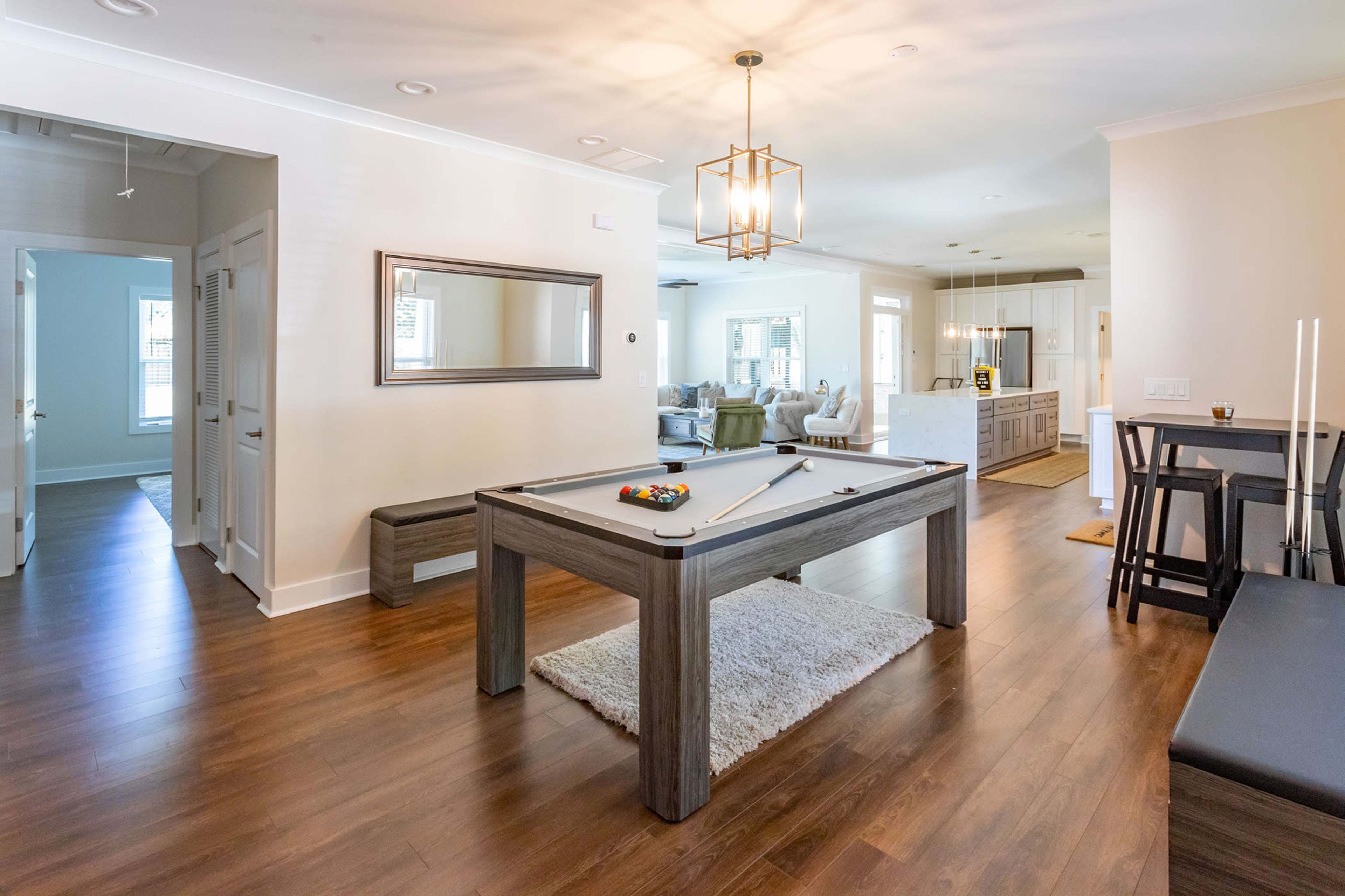 A modern living space featuring a pool table in the foreground, a dining area with high stools, and an open kitchen in the background.