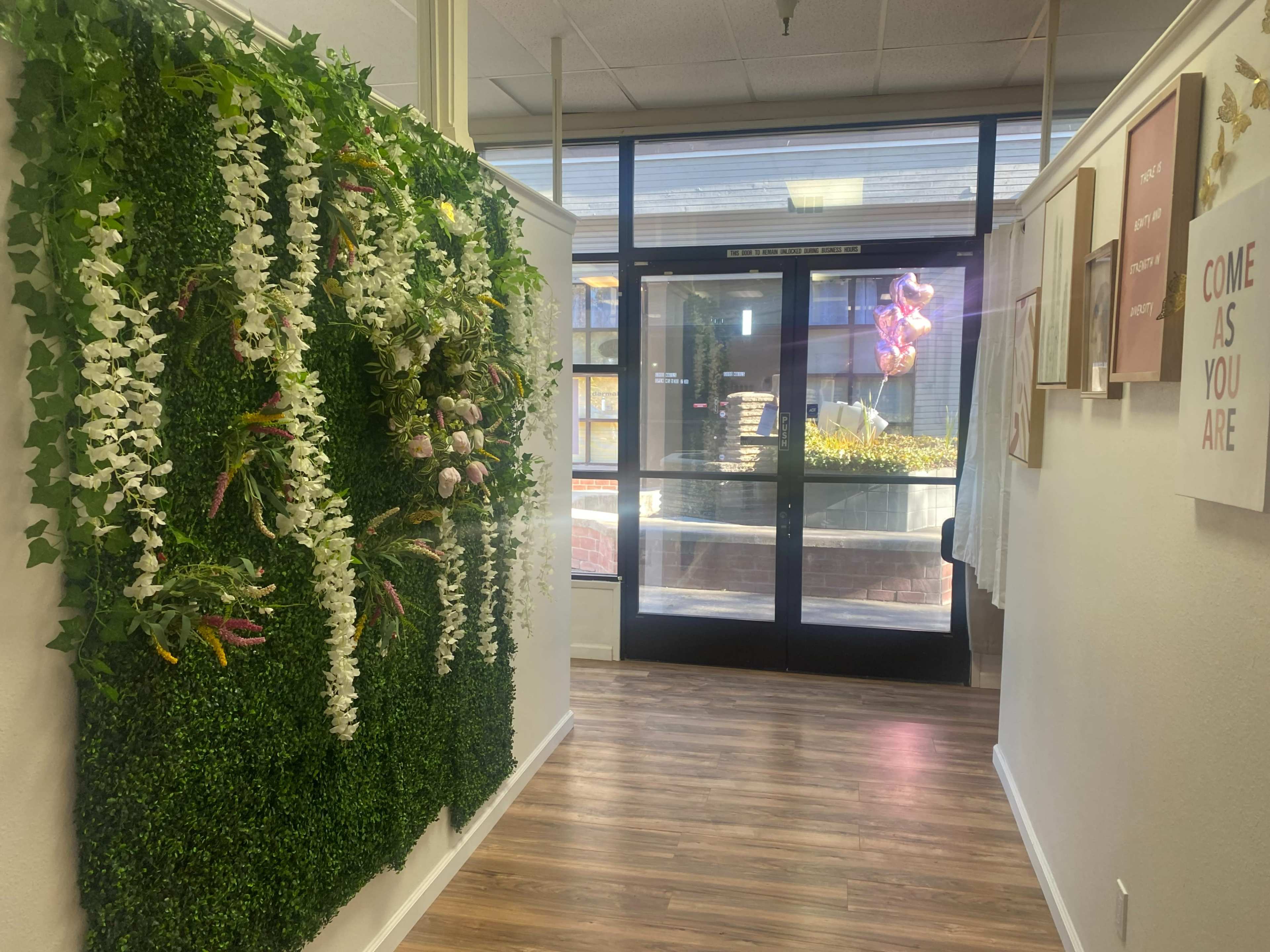 A hallway with a decorative wall of greenery and flowers leads to a glass entrance door.
