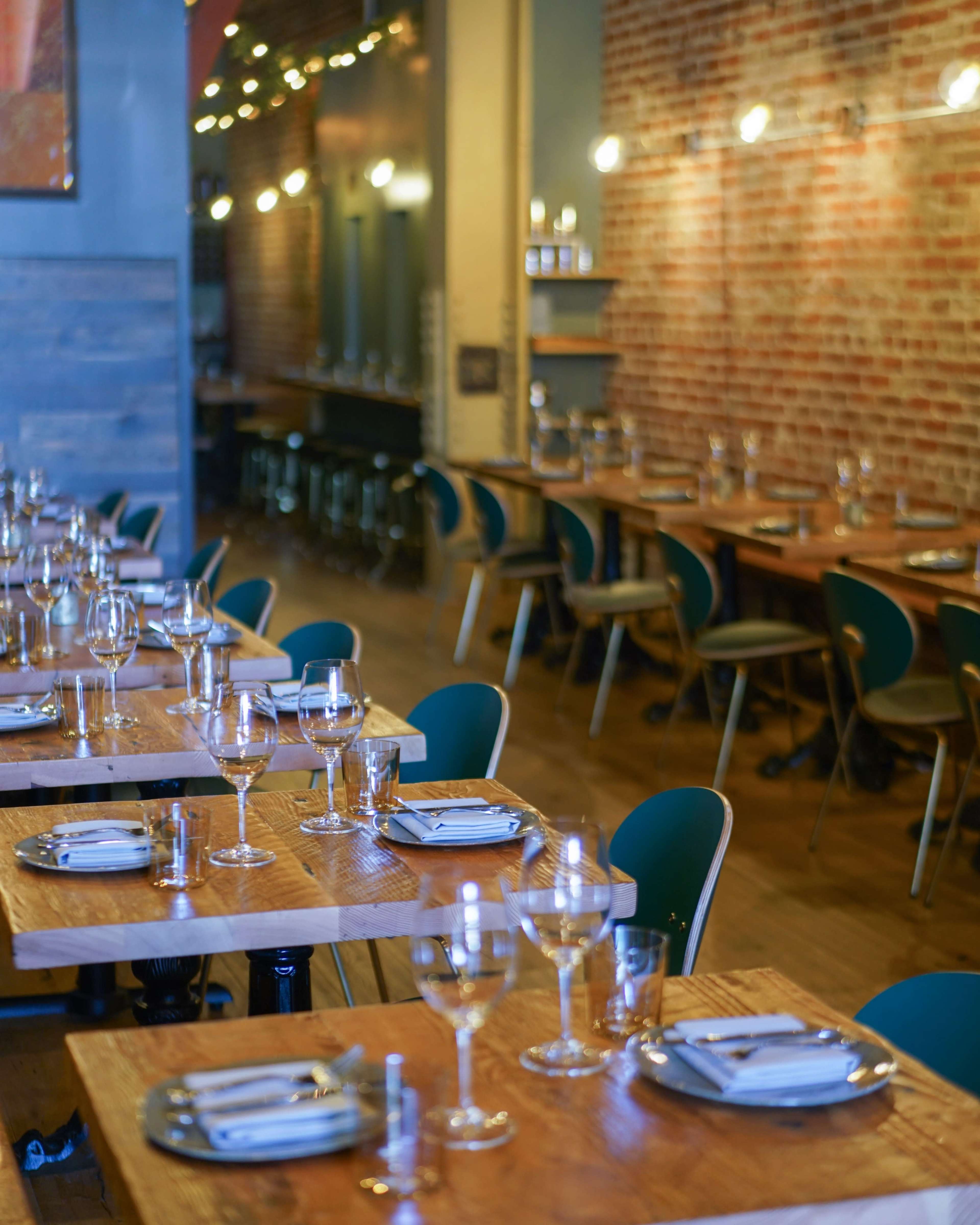 The image shows a restaurant interior with long, wooden tables set for dining, featuring glassware and cutlery, against a backdrop of exposed brick walls and minimalist decor.