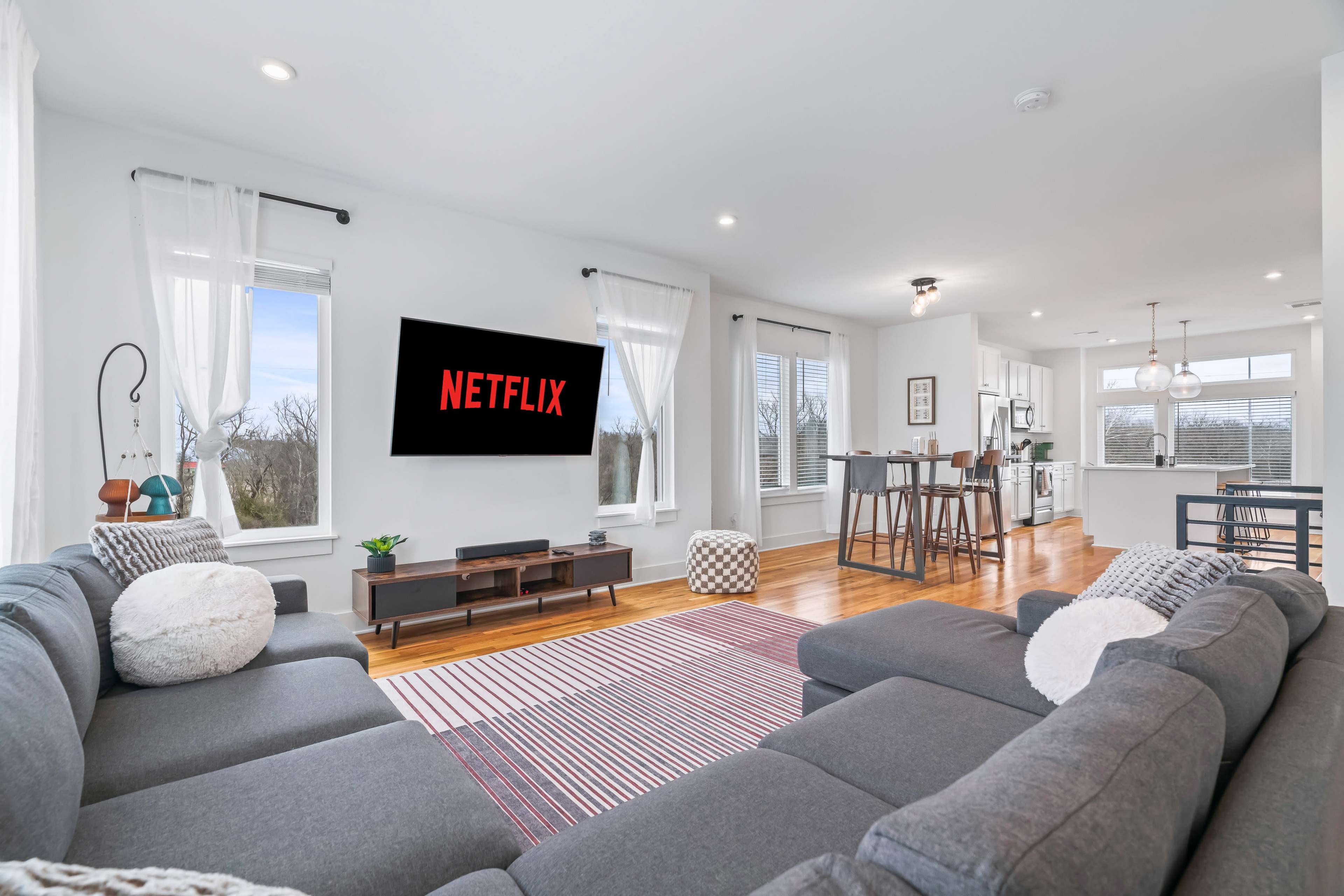 A modern living room features a large sectional sofa, a wall-mounted TV displaying the Netflix logo, and an open kitchen in the background.