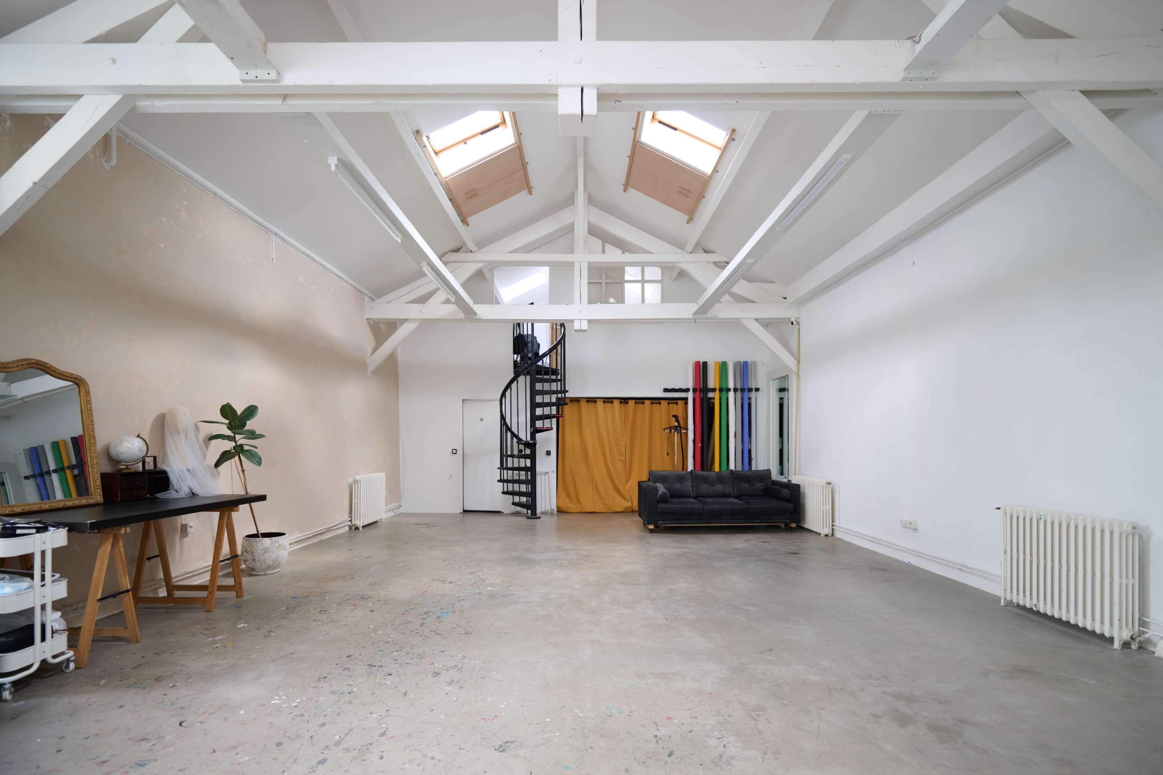 The image shows a spacious, minimalist studio with high ceilings, wooden beams, and a spiral staircase, featuring a black couch, a desk, and colorful vertical panels against the wall.